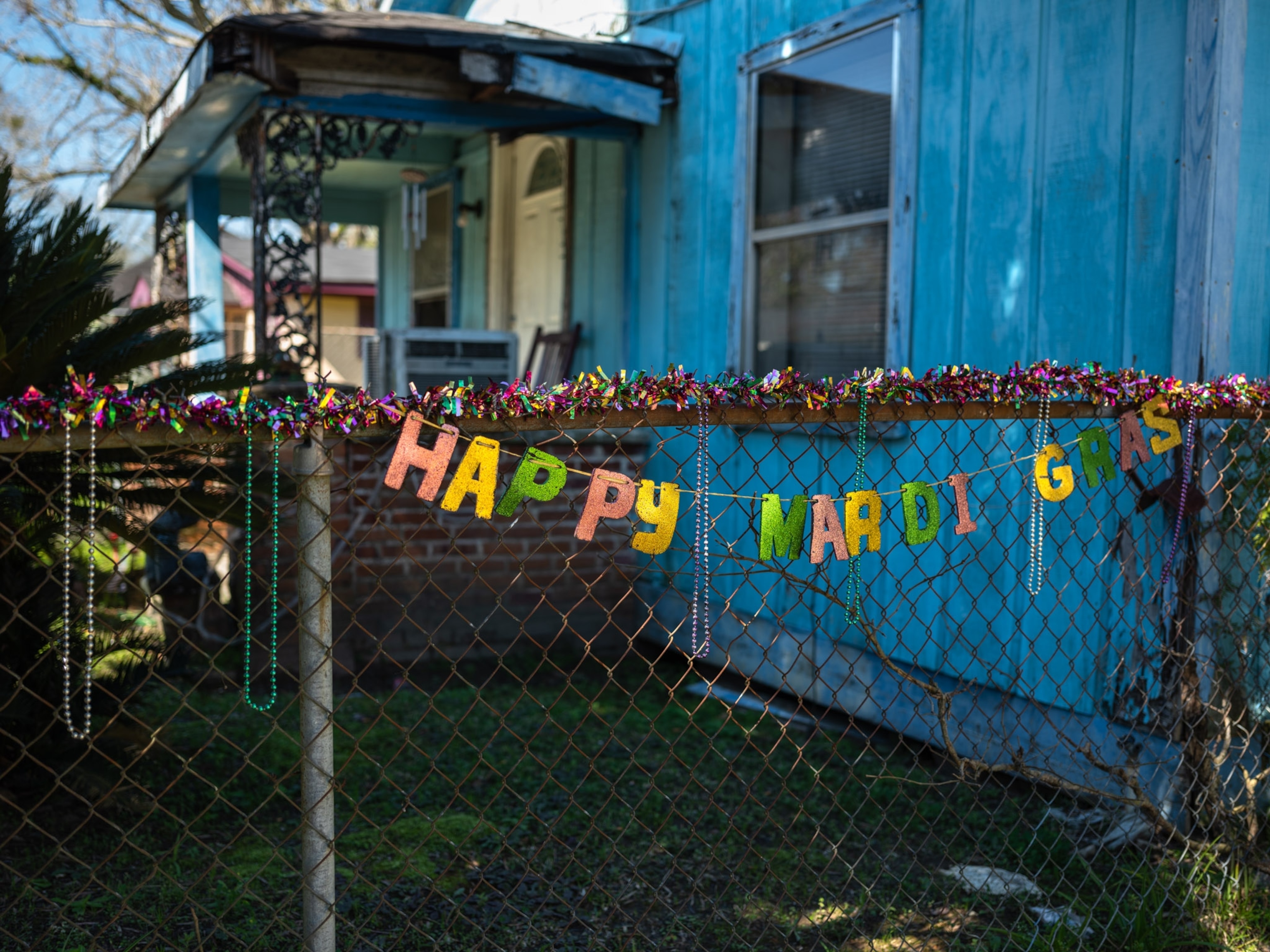 a hanging lettered sign on a fence that states "happy mardi gras"