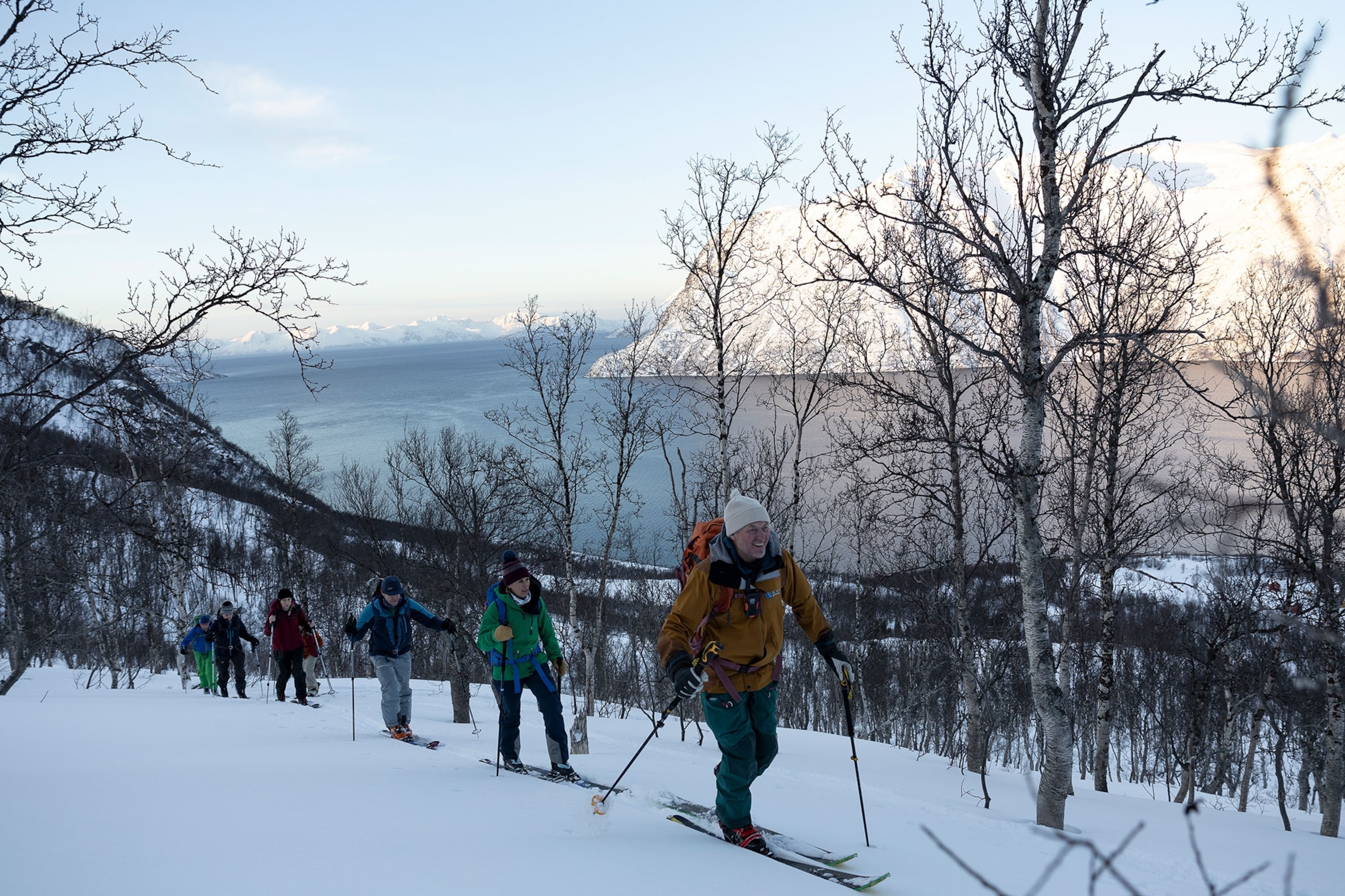 A group of skiers make their way across a snow covered path surrounded by trees. In the background is a body of water surrounded by mountains.