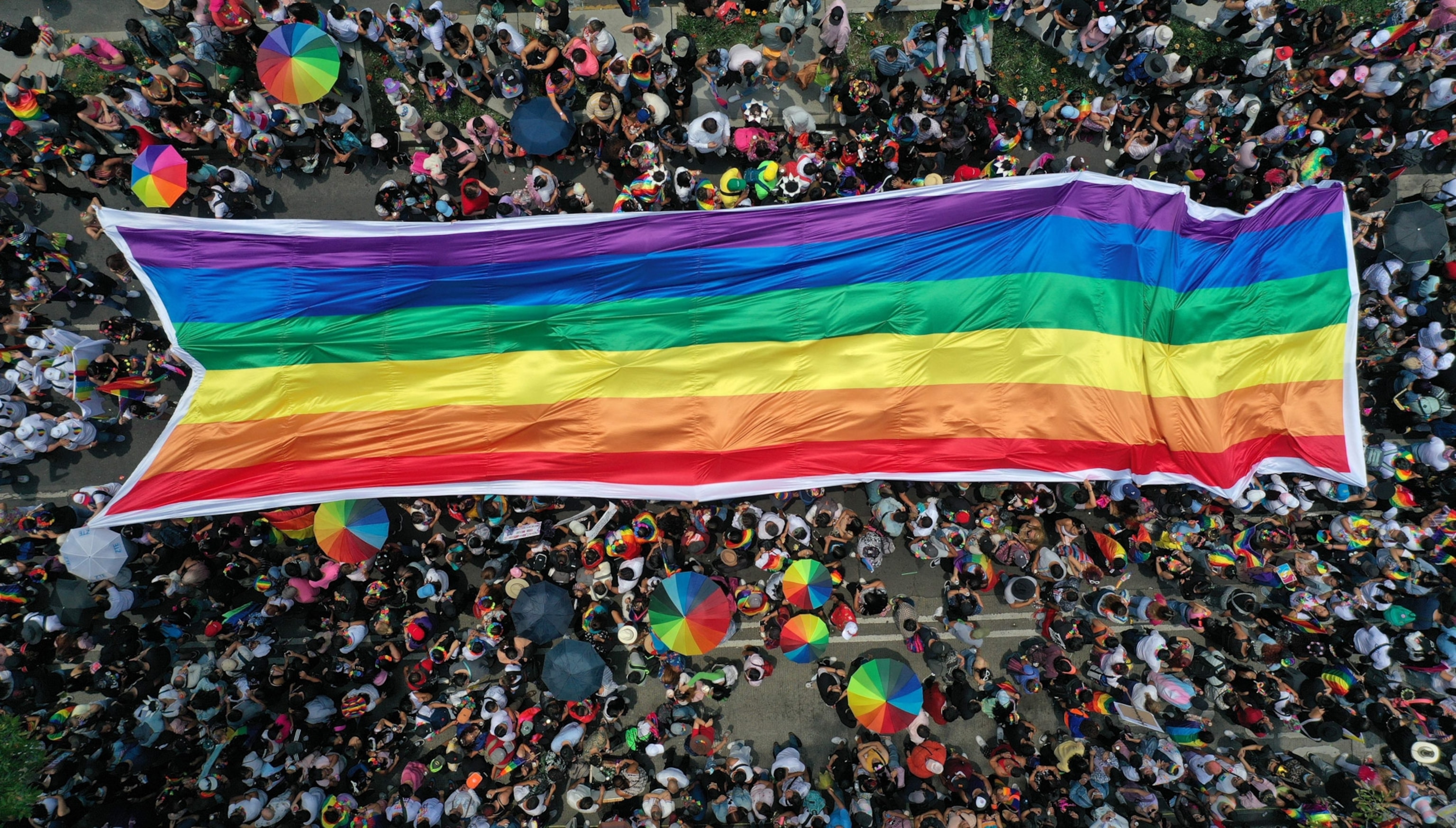 A top view of people in a pride parade with a large pride in the center and people walking with pride umbrellas around the flag. It's a densely packed parade.