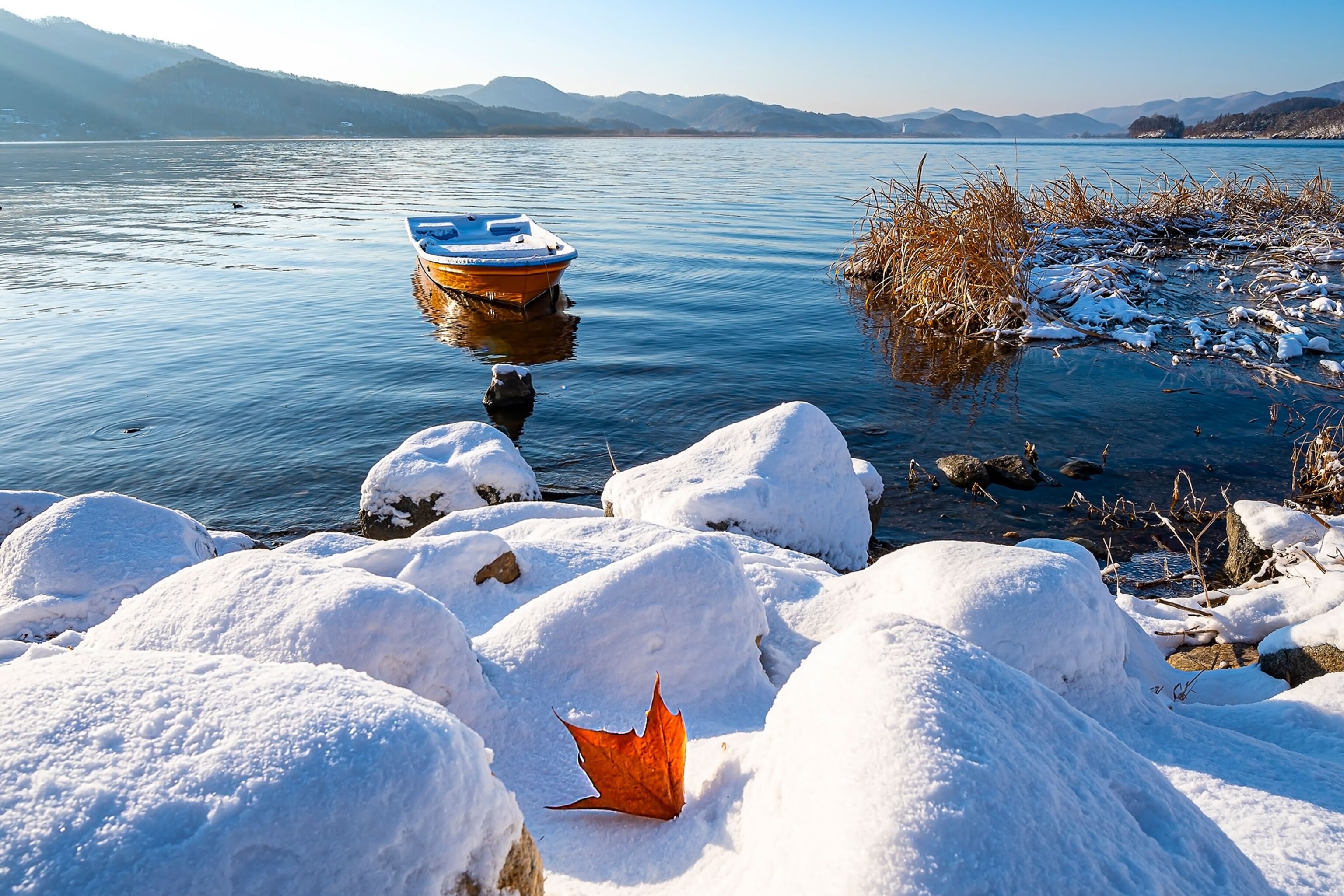 A rowboat is seen on a calm, blue lake, with snow-covered shoes in the foreground.