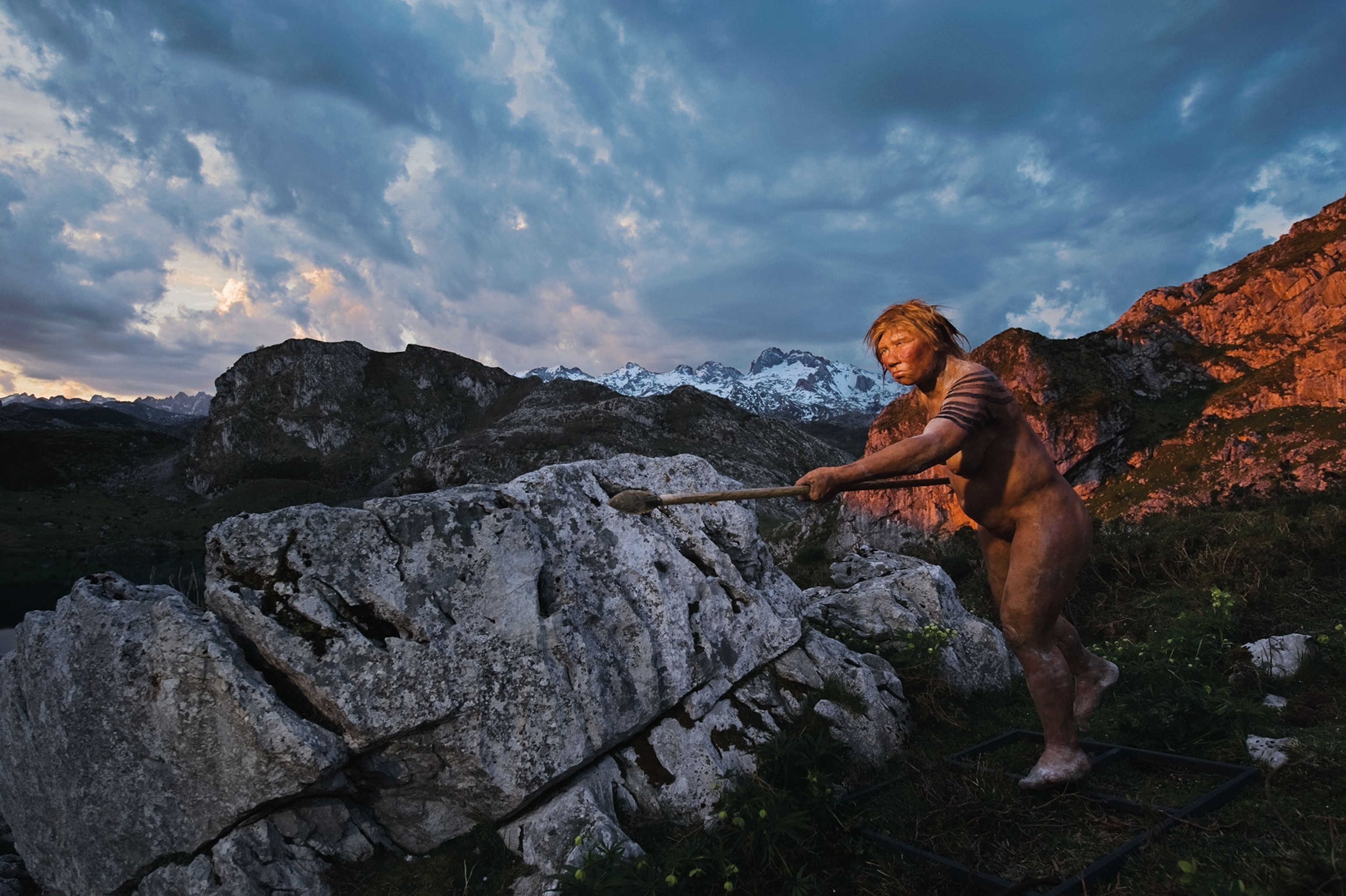A reconstruction of a Neanderthal female in furs stands on a mountain holding a spear