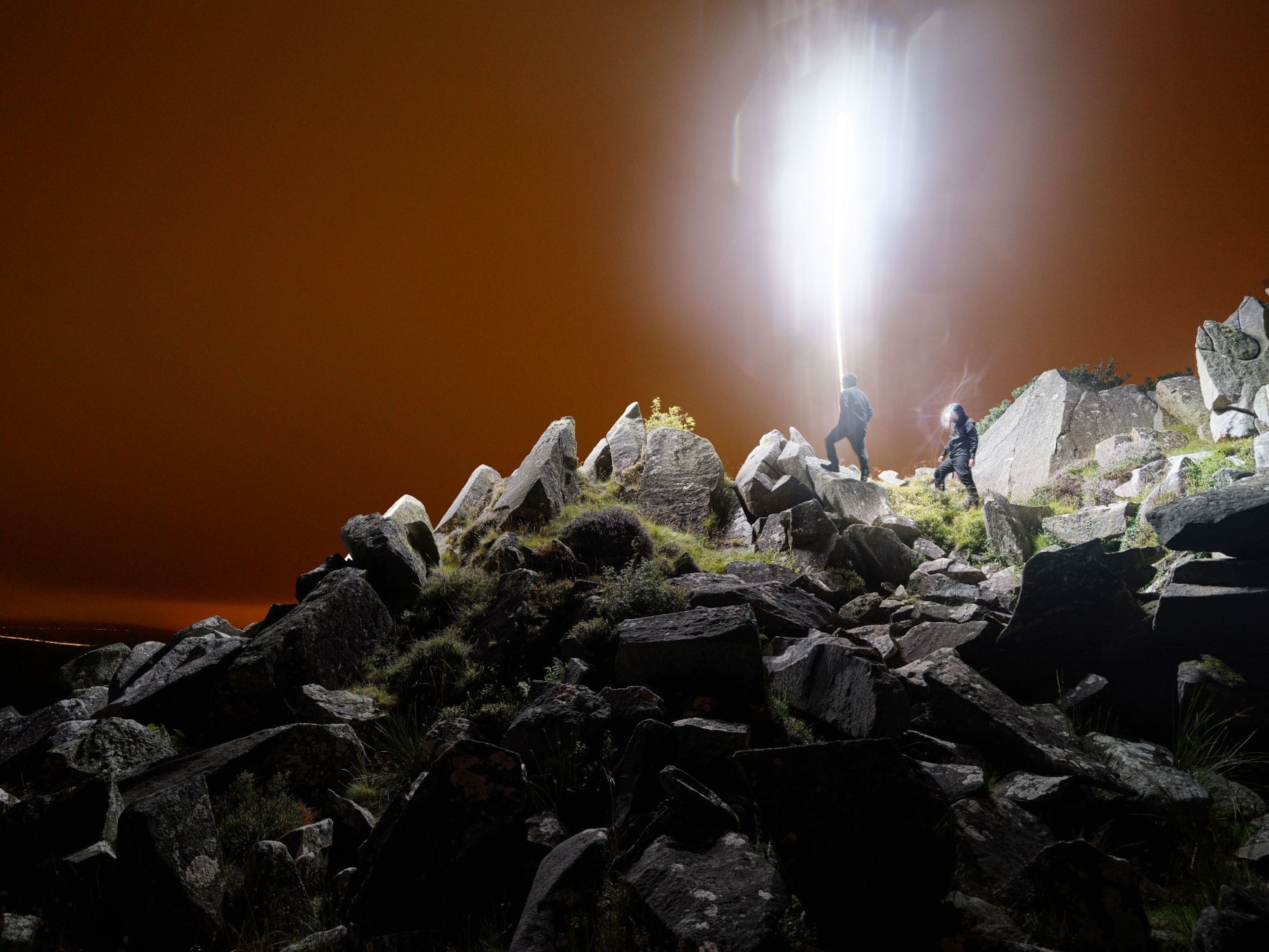 Two men stand on a rocky outcrop illuminated by a drone