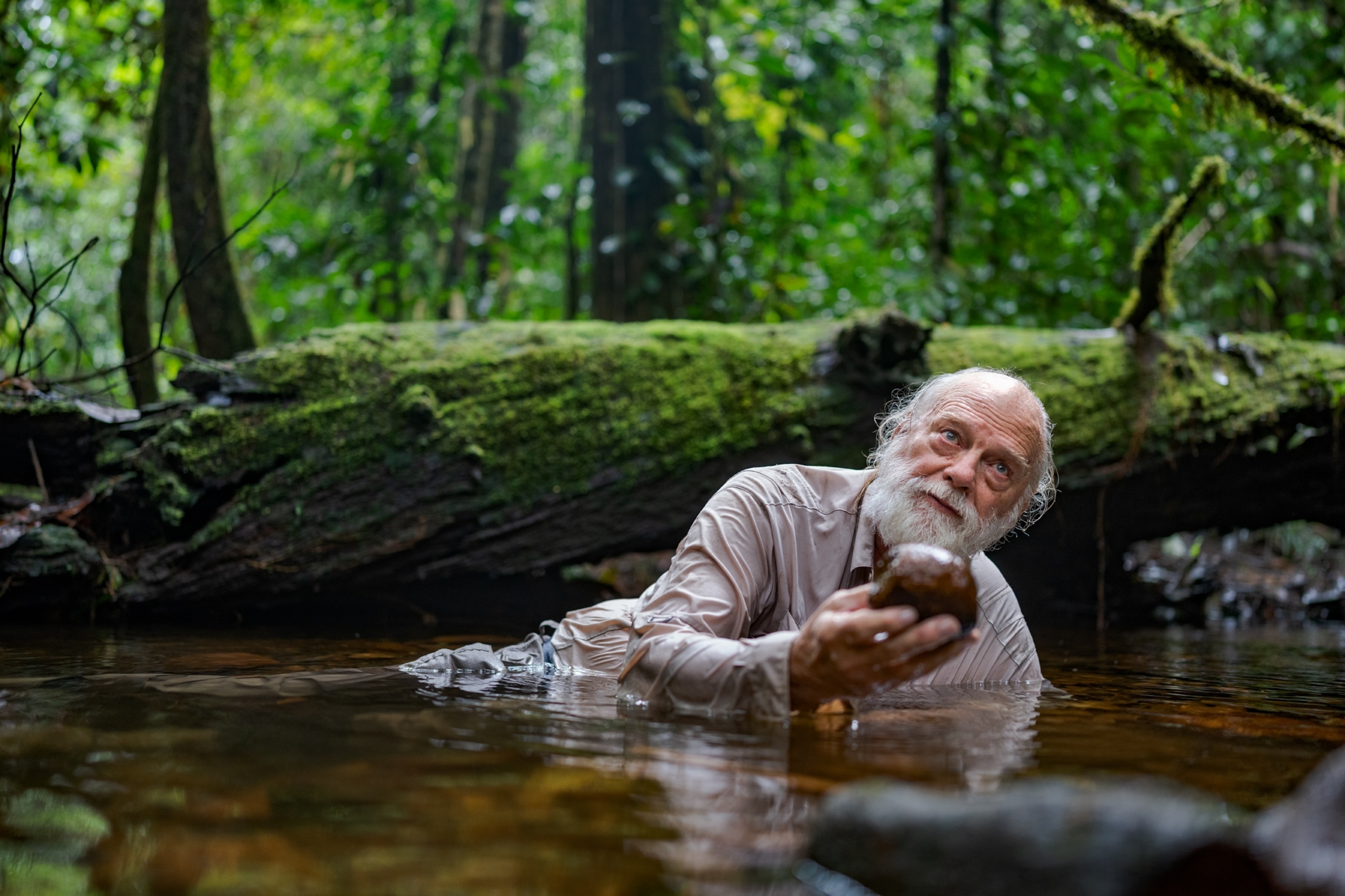 Picture of man with gray hair lying in water and showing round rock in his stretched out hand.