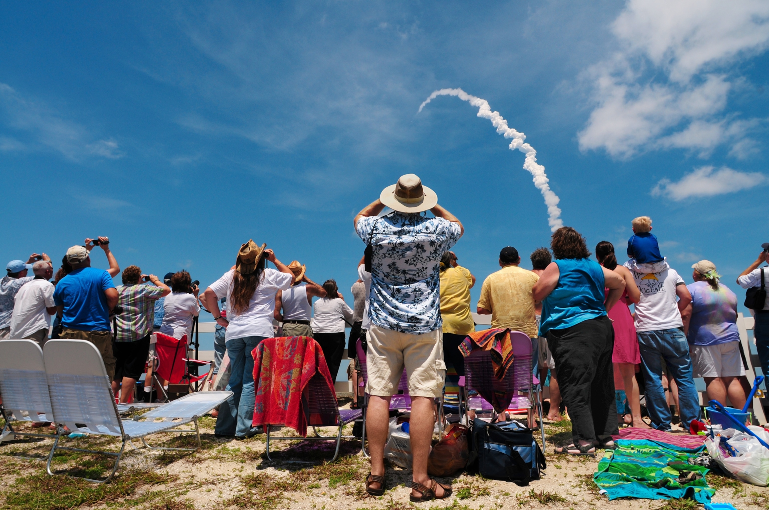 A large group of people dressed in warm weather clothing stand and sit along a beach as the cloudy jet trail of a launched rocket arcs through the sky.