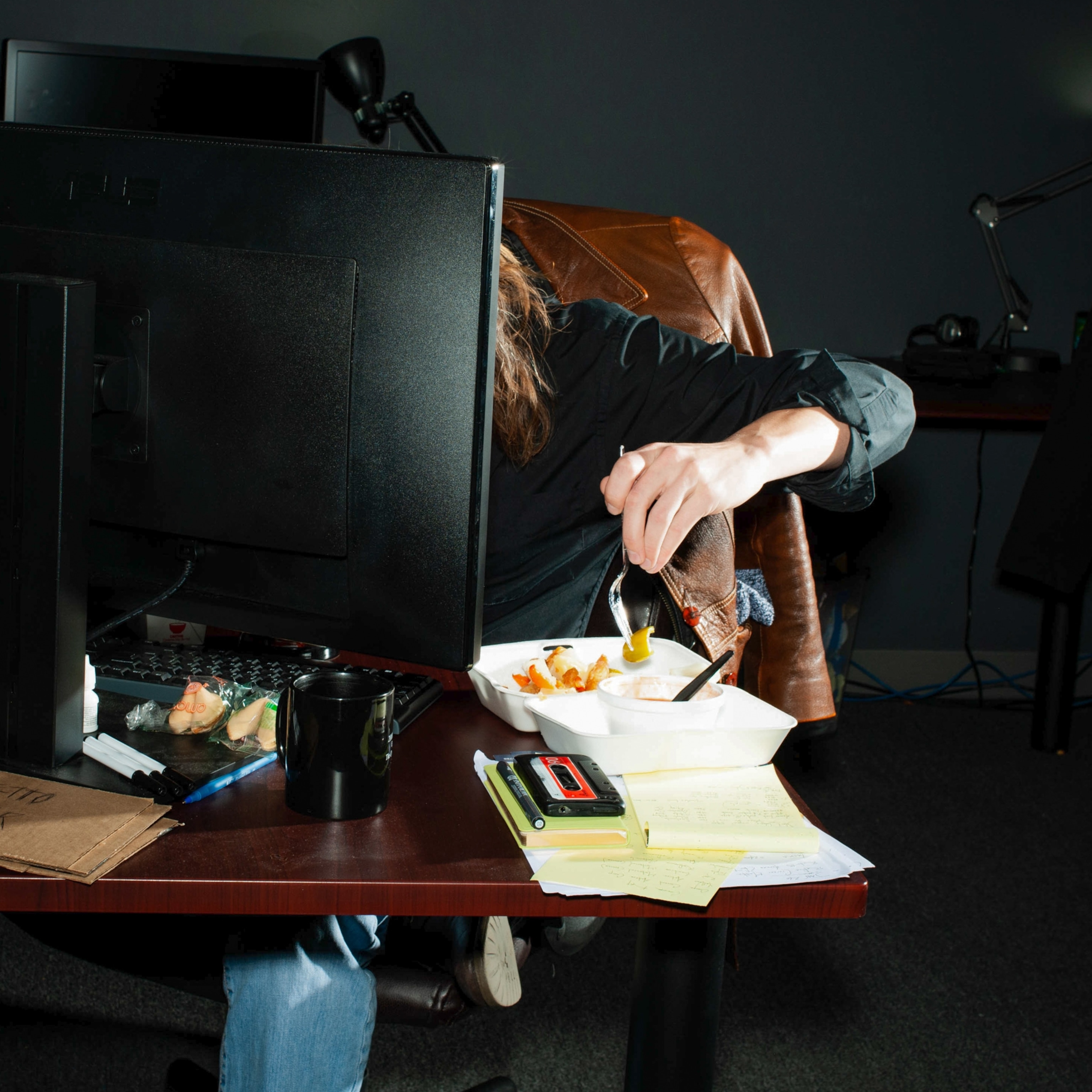 A computer monitor blocks the face of someone as they eat Chinese take-out at their desk.