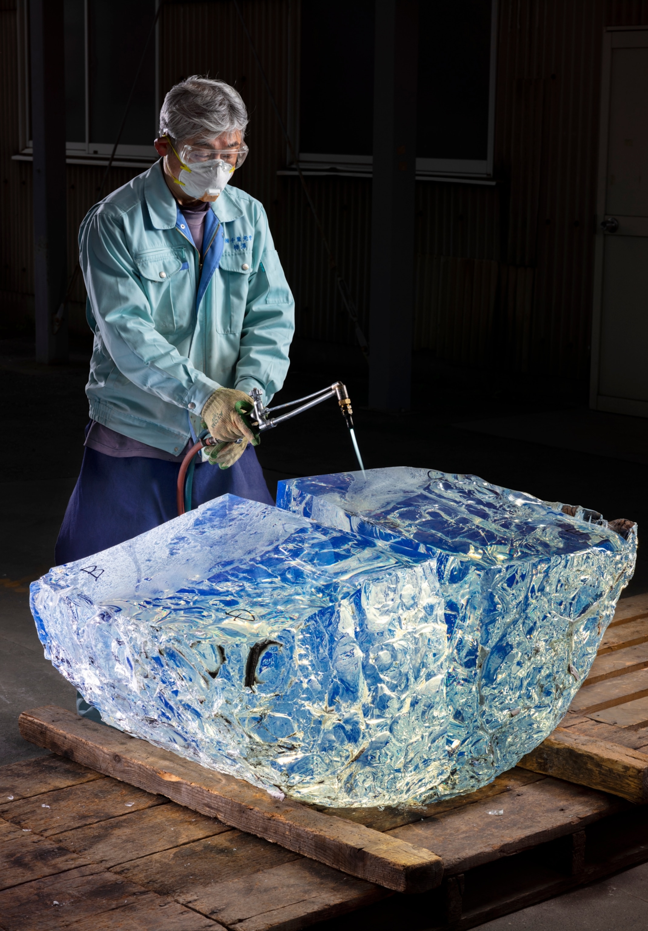 a man in aa blue shirt holding a torch over a large unshaped for of glass as it splits down the center.