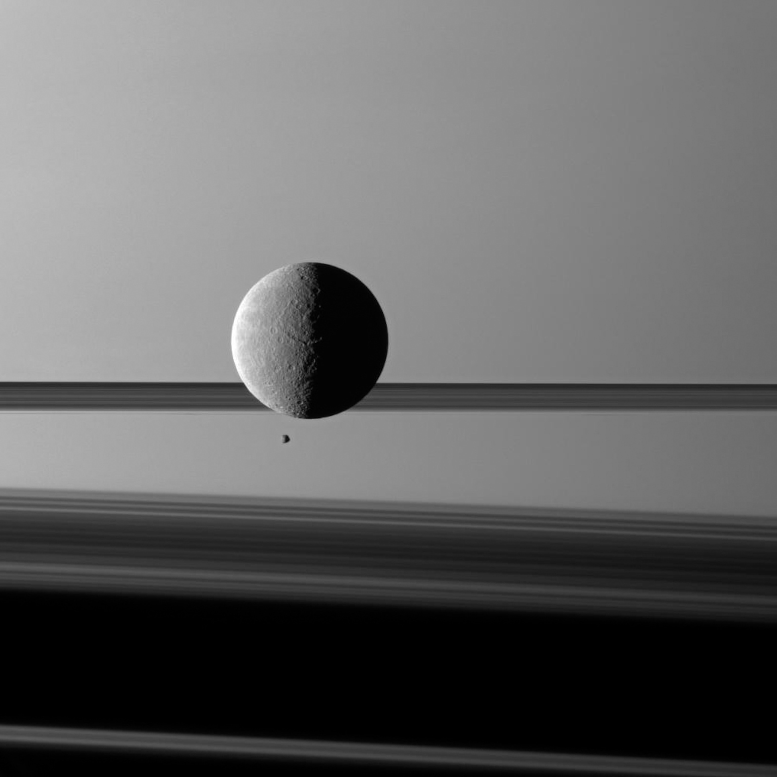 A large round moon seems gigantic as it looms over a tiny rocky moon, against the backdrop of bands of Saturn's rings.