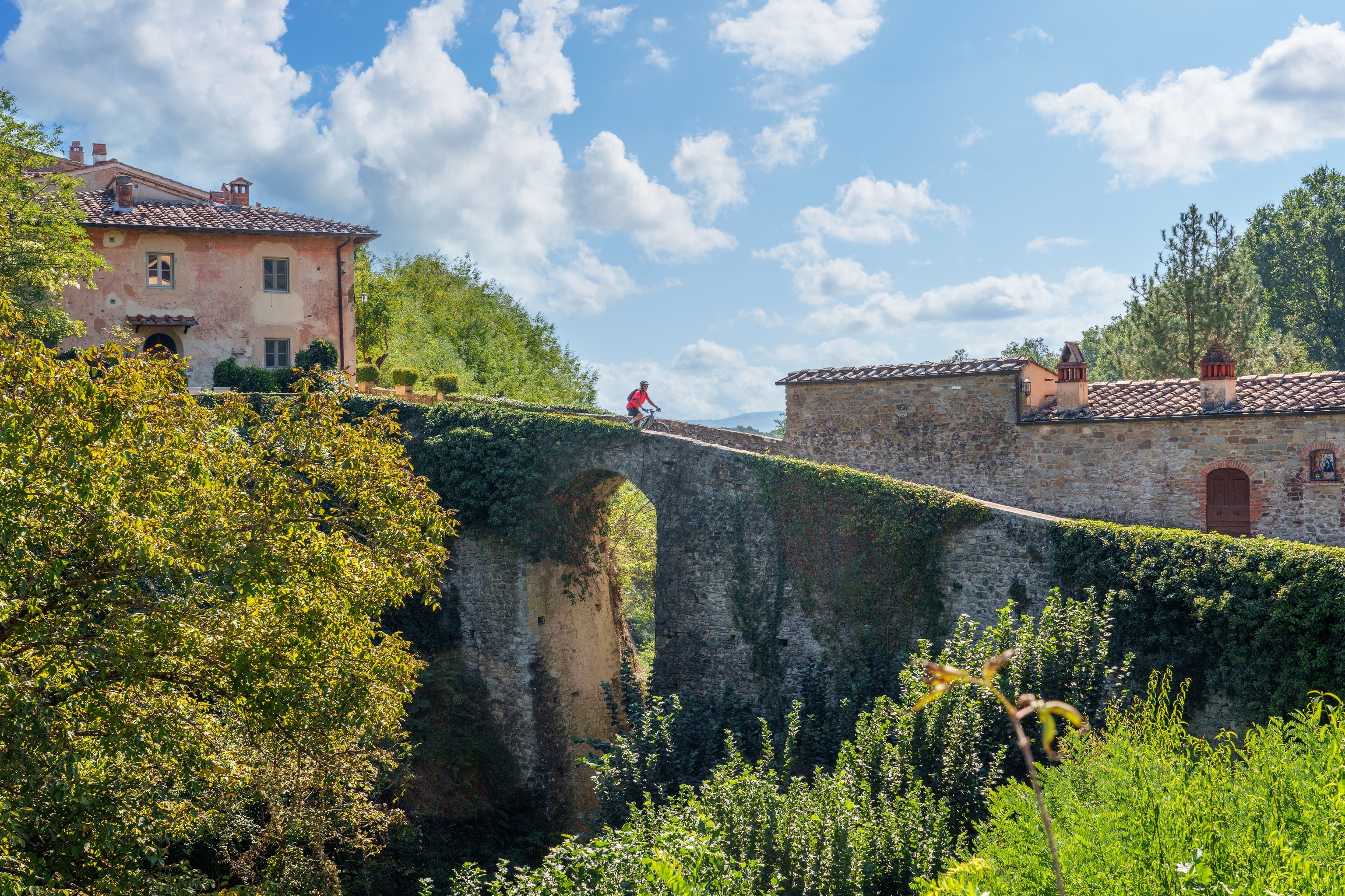 A woman wearing a bright red shirt, is cycling over an old, stone bridge, in a Tuscan village with brick houses with terracotta shingles.