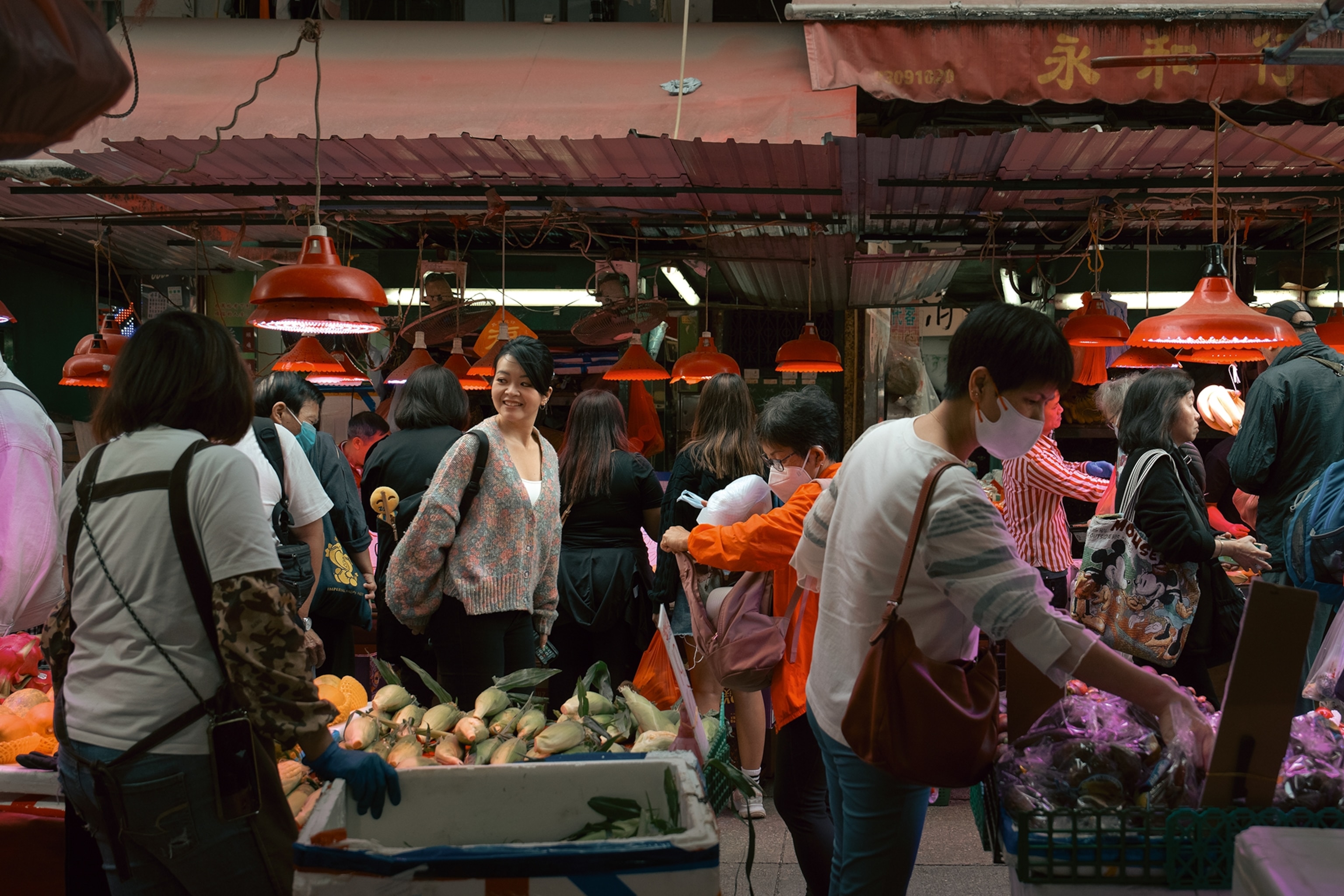 A woman strolling over a busy, half-roofed market with vendors selling vegetables.