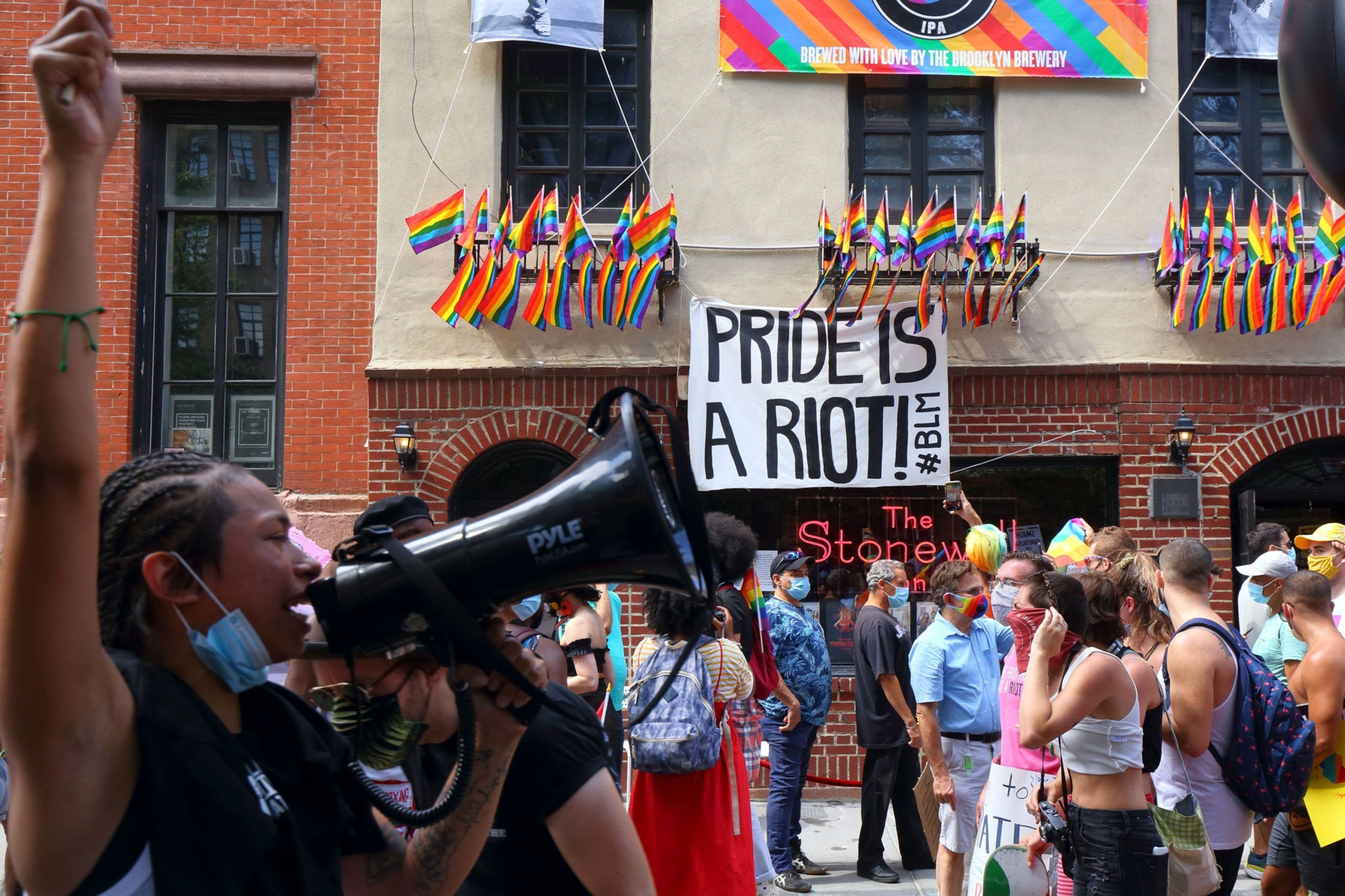 a crowd gathers outside Stonewall in in New York. Rainbow pride flags are hanging off the building along with a sign that says "pride is a riot"