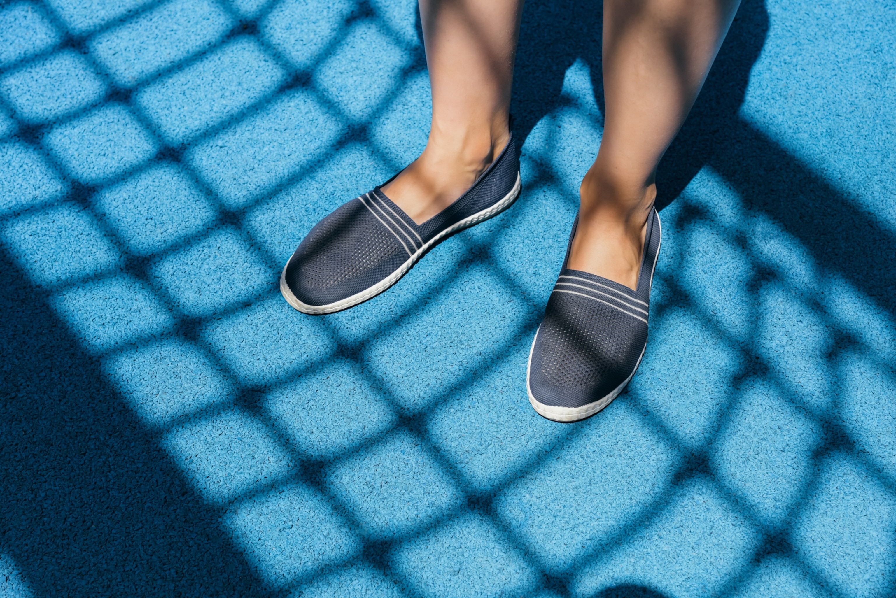 The lower legs of a man wearing blue slip-on shoes on a blue tennis court. The shadow of the tennis net casts a pattern over the ground as well as the man's legs and shoes.