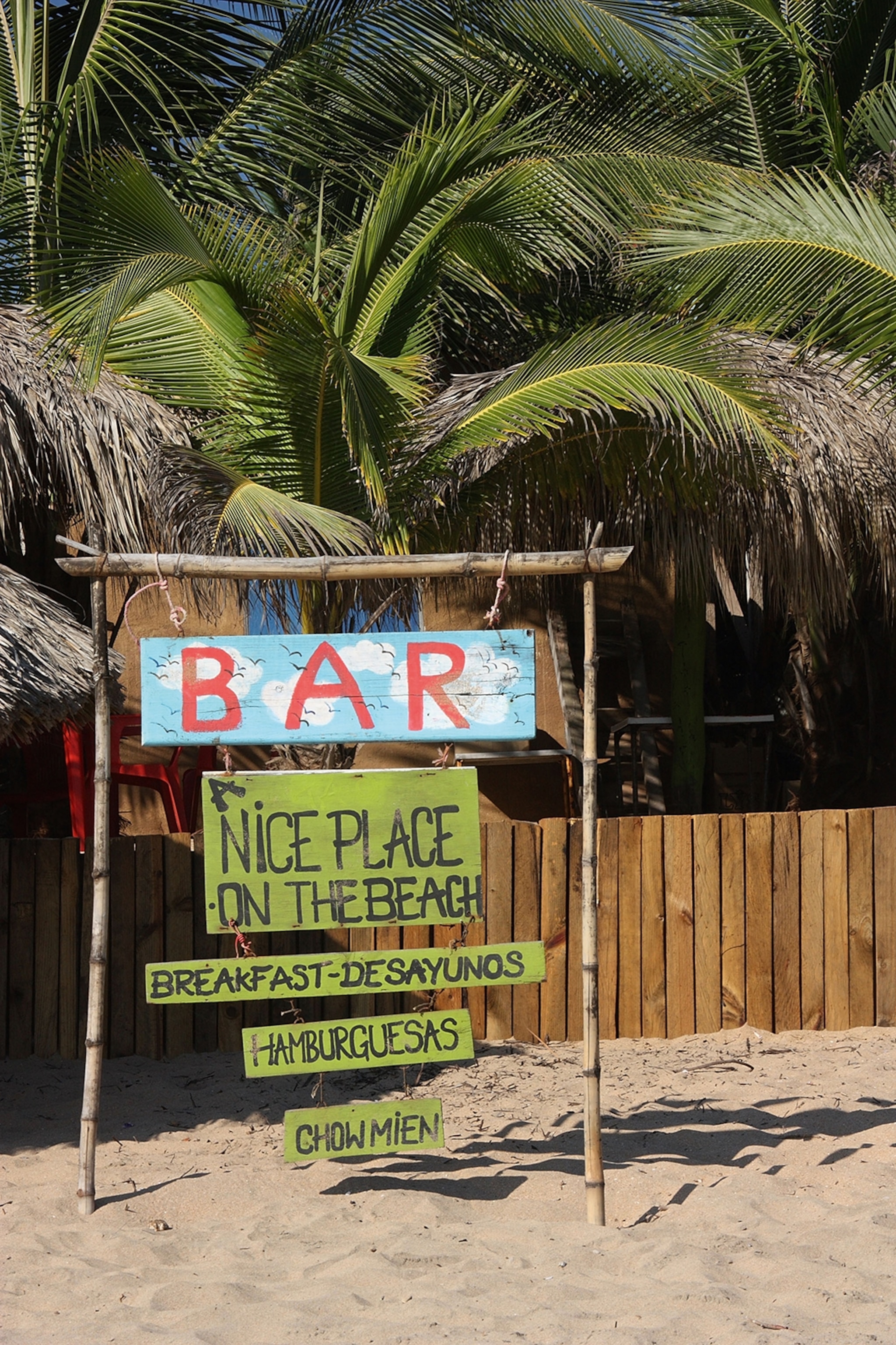 A sign on a beach inviting to a bar with palm trees in the back.