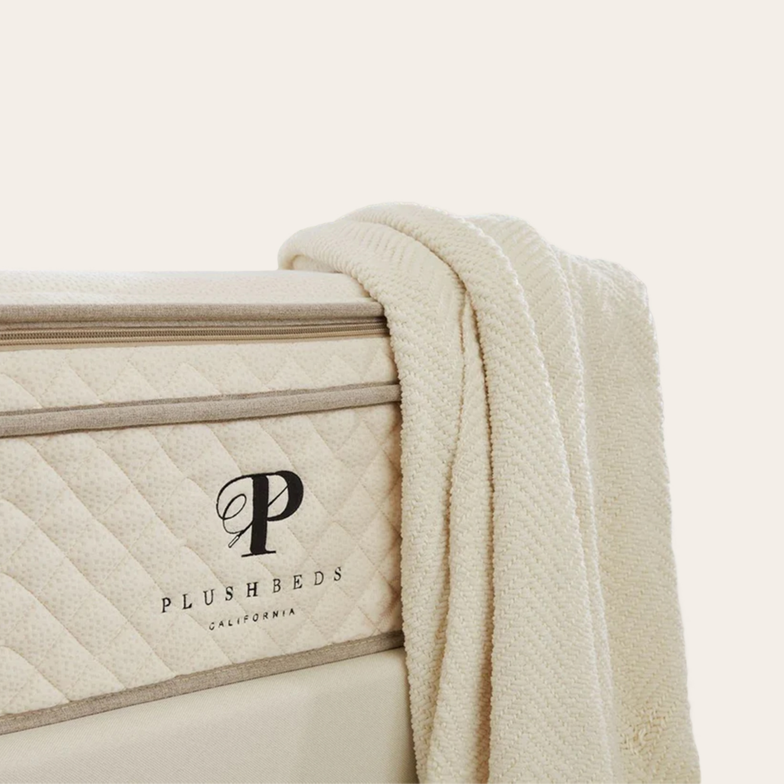 A close-up view of the corner of a mattress with a blanket draped over the side; against a tan background.