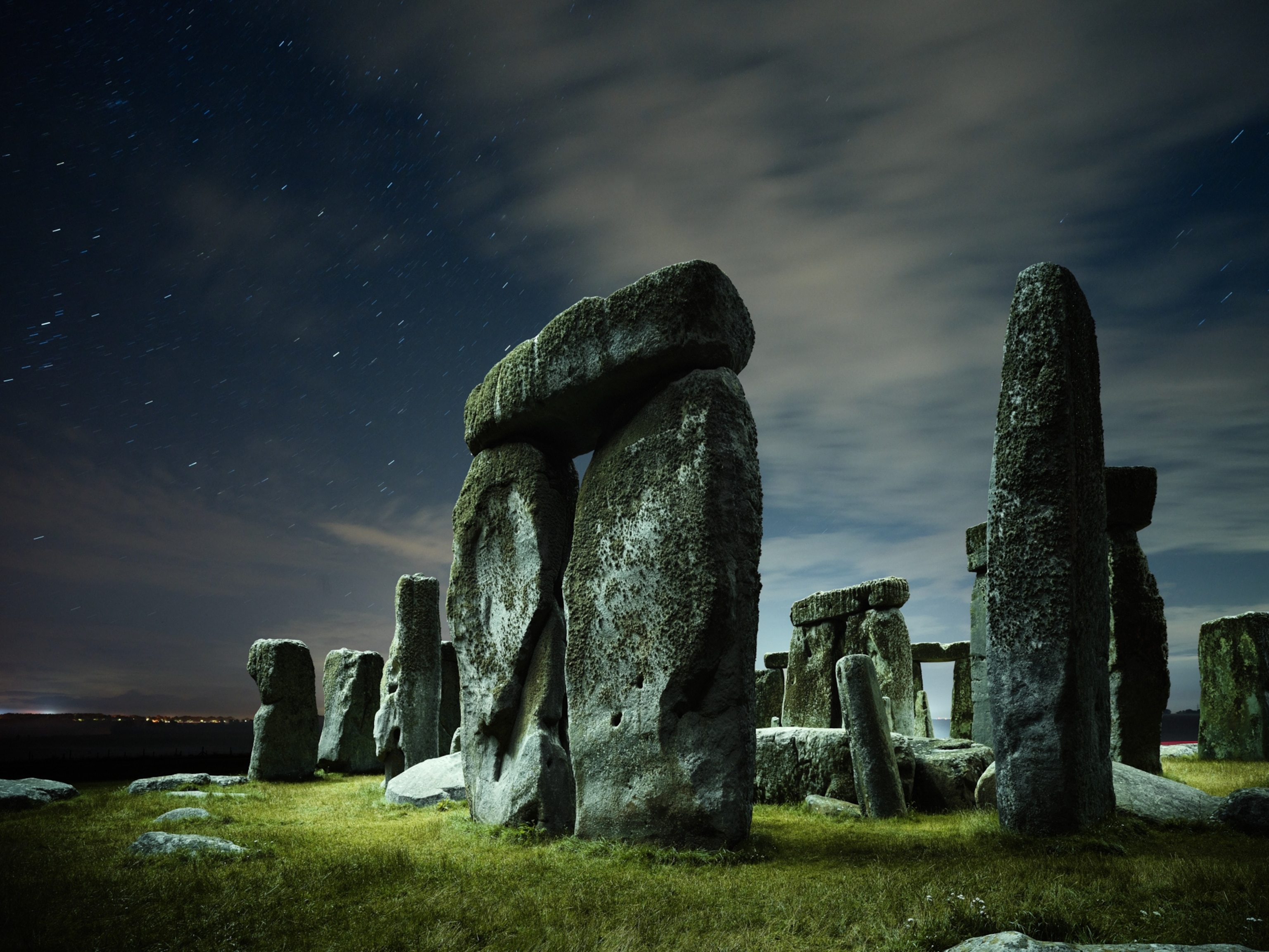 A close-up, wide angle view of some of the standing stones of Stonehenge, seen beneath a partly cloudy and starry sky at night.