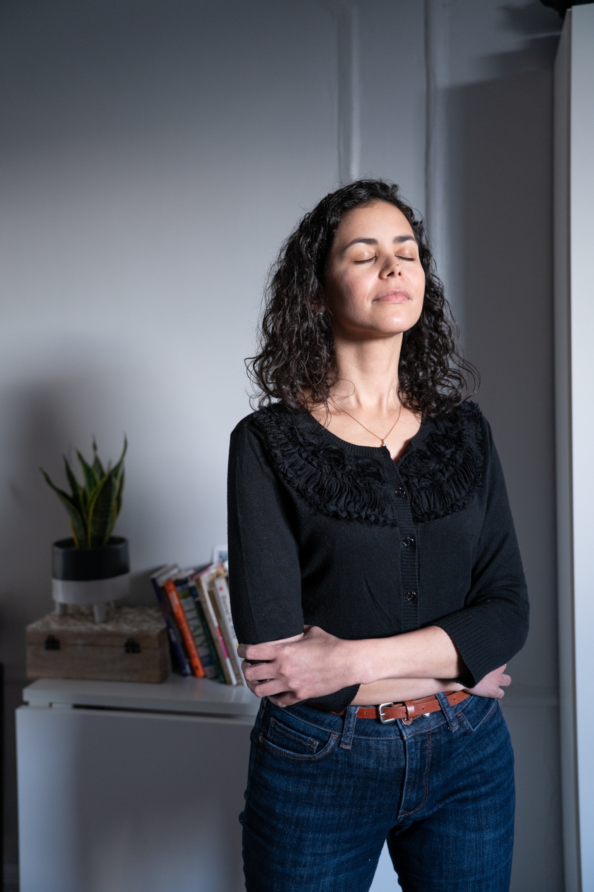 Aline embraces herself as she stands in the sunlight filtering in through her window.