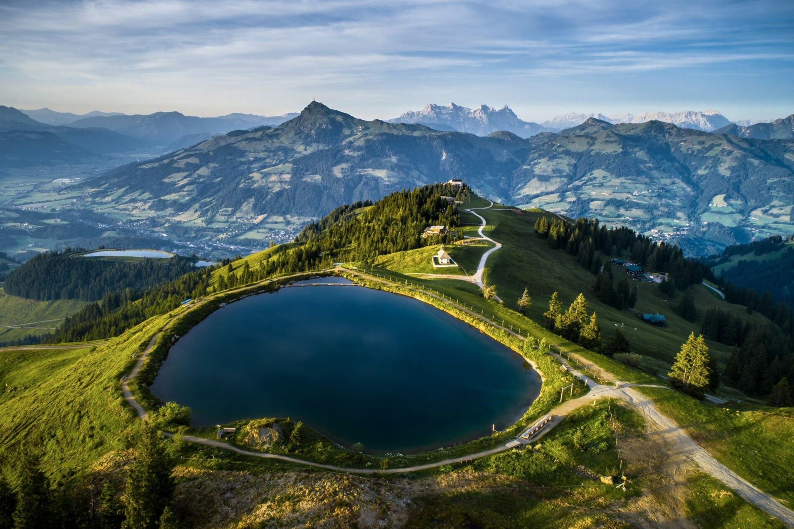 A circular pool of water, atop a green hill, in front of a long valley populated by small buildings.