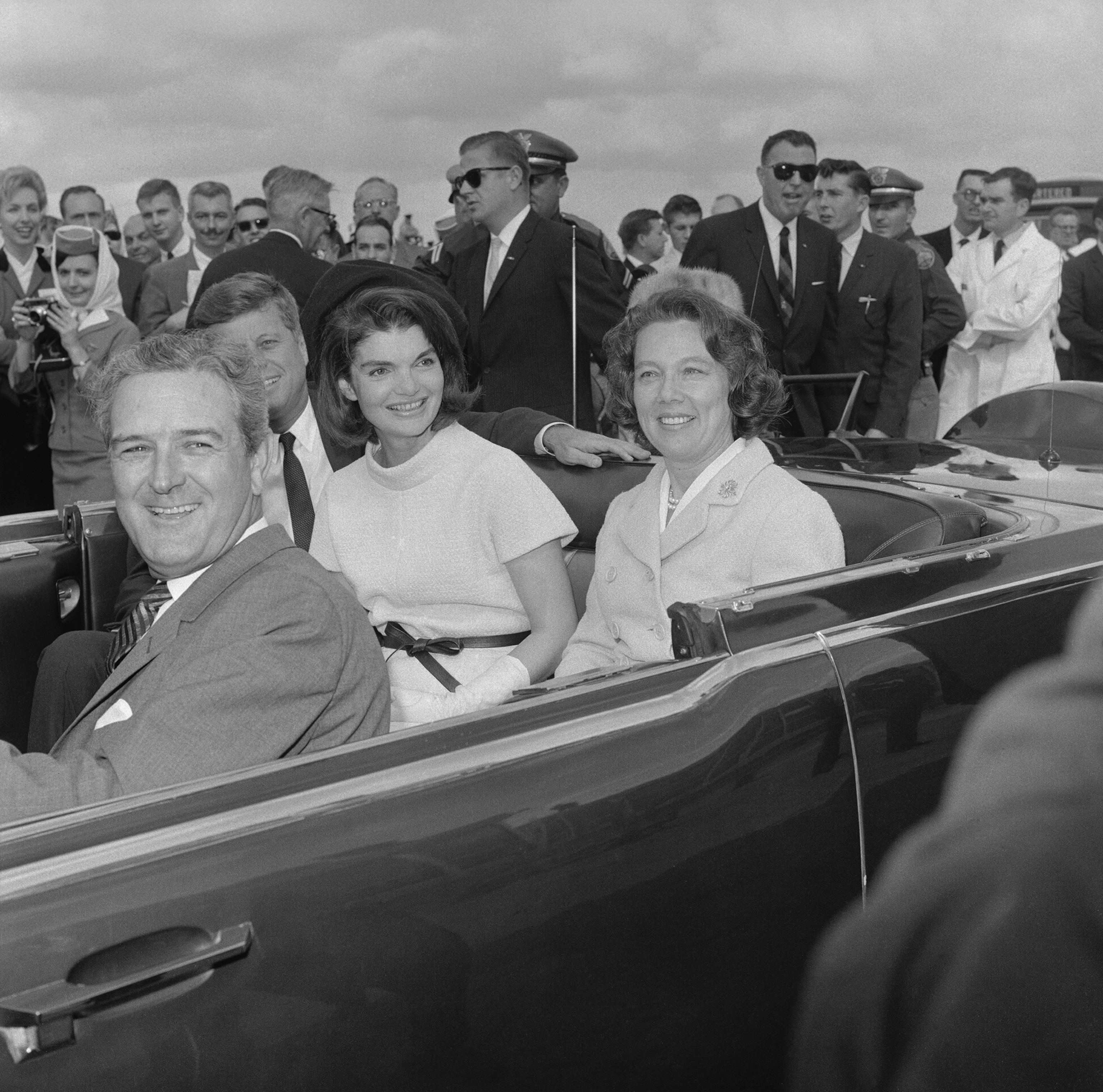 President Kennedy and First Lady Jacqueline Kennedy sit next to Texas Governor and Mrs. Connally in a car surrounded by a crowd..