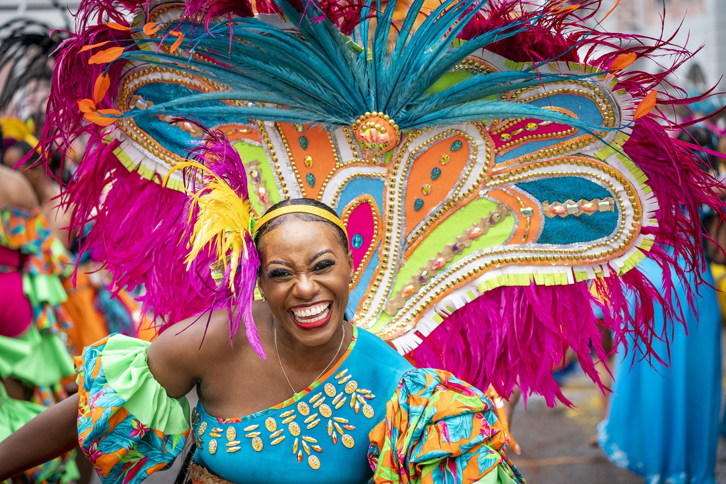 A woman dressed in bright, multi-colored clothing, with a large, multicolored and multi-textured peacock-like costume tail dances in the street with other performers.