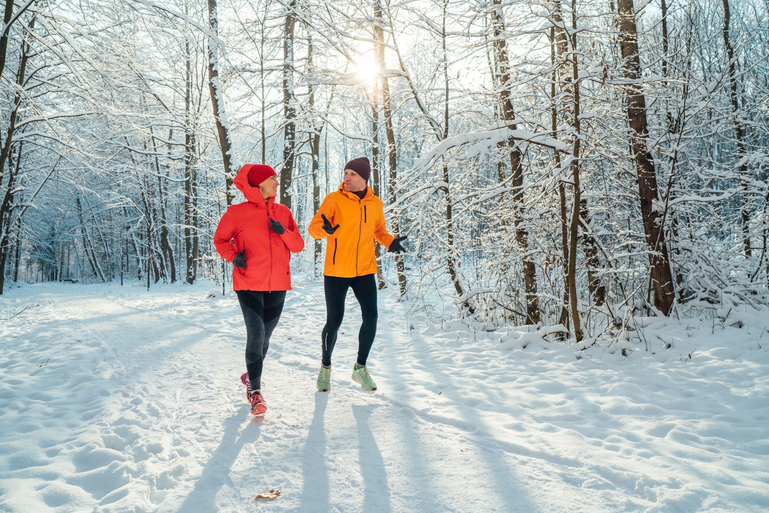 A middle-aged man and woman running along a snowy woodland trail with sun shining in through the trees from behind.