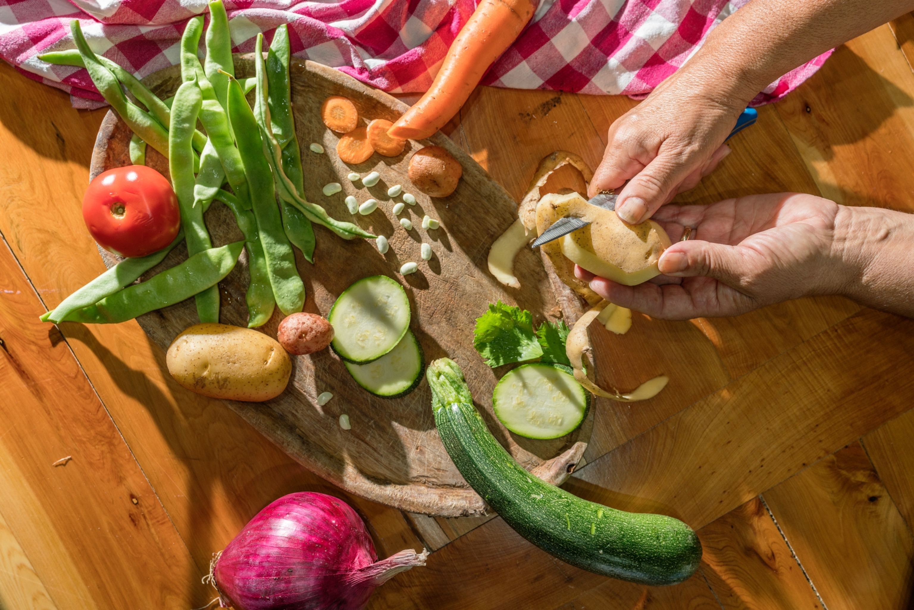 An overhead view of hands peeling a potato over a cutting board covered with various vegetables.