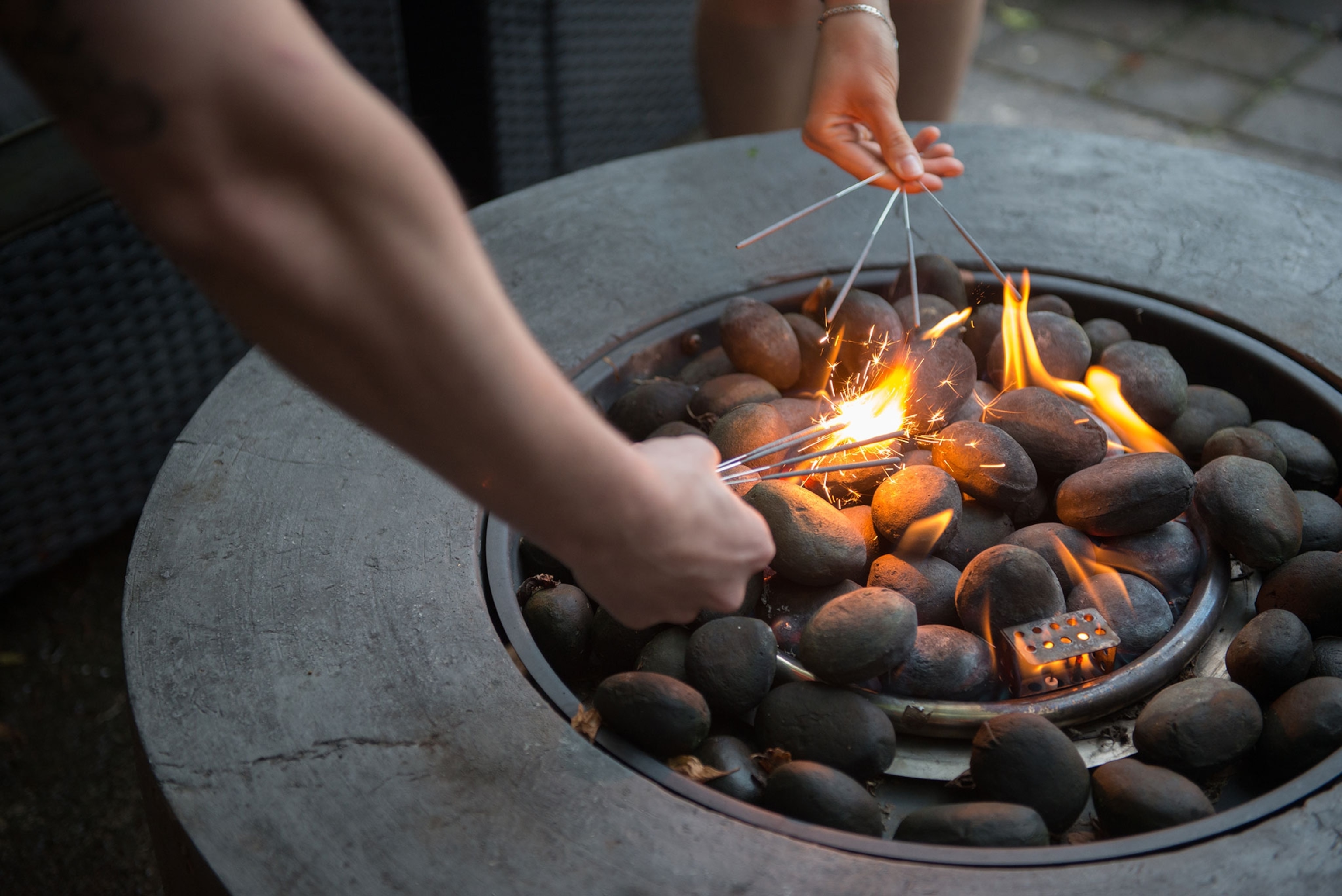 Two people reach arms over top a round grey fire pit to light sparklers.