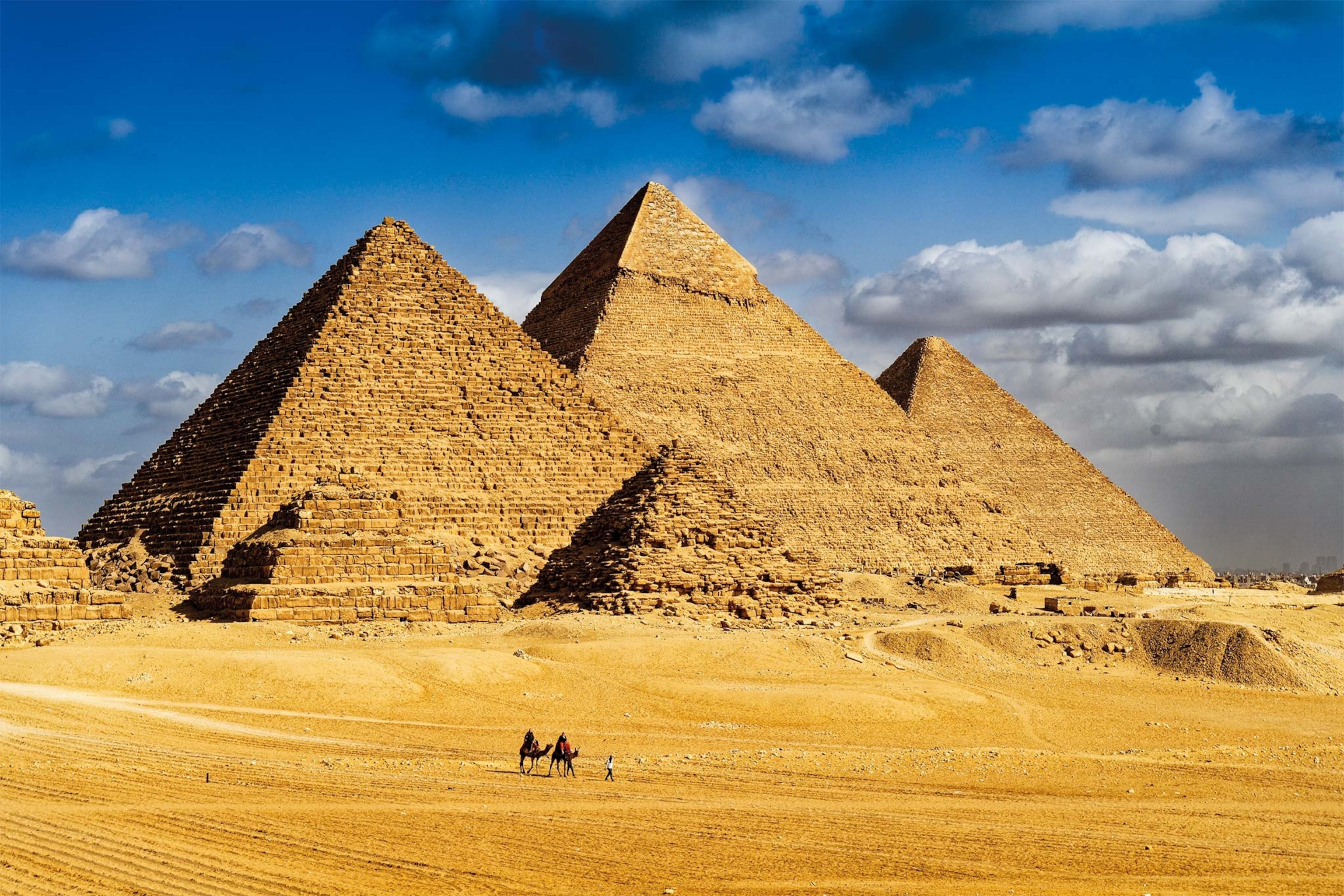 A view of the Pyramids of Giza, with blue sky behind, sand in front, and two camels in the foreground