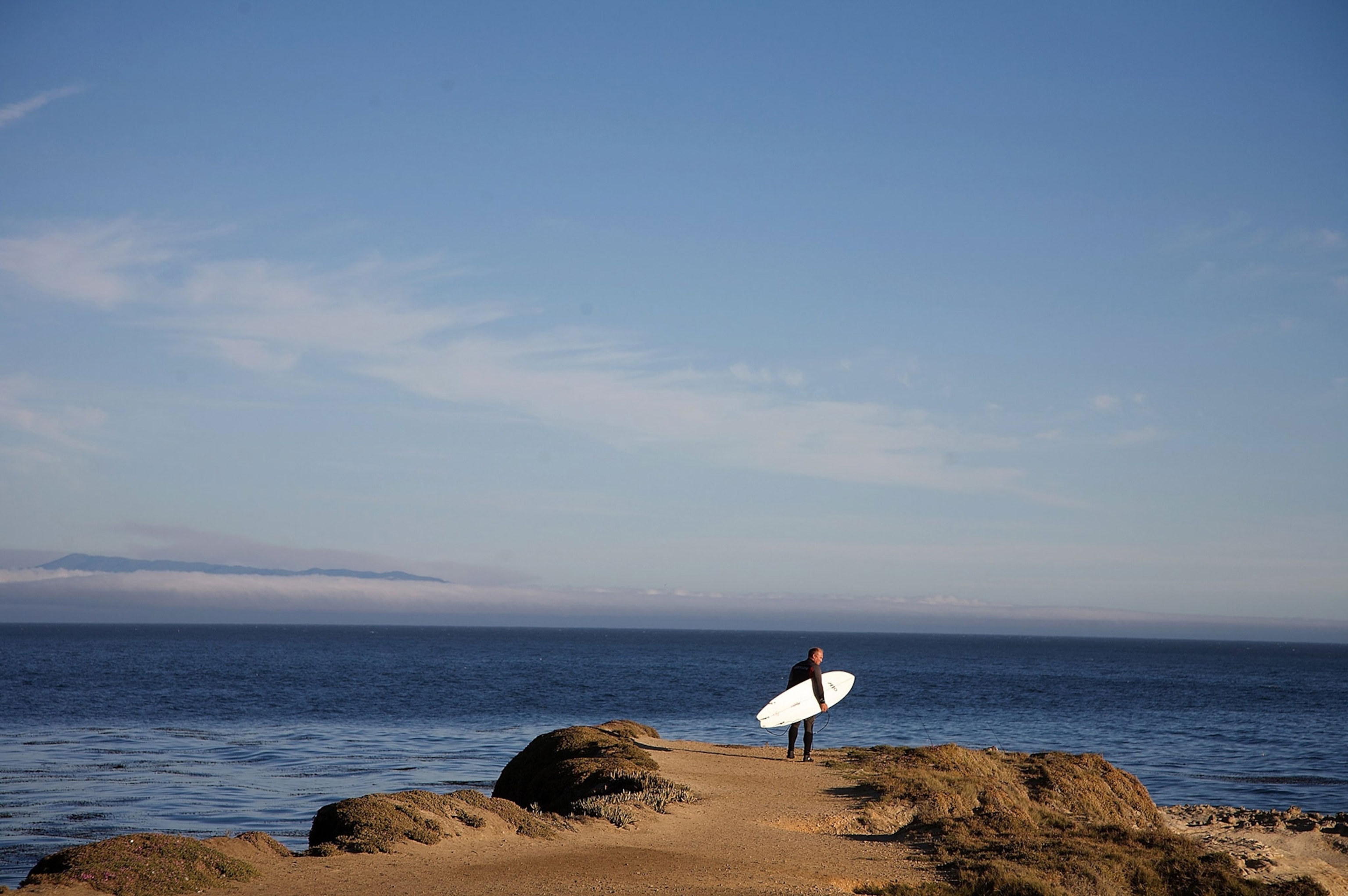 A surfer stands towards the edge of a cliff looking off to the distance at the ocean while holding a white surf board in their arms.