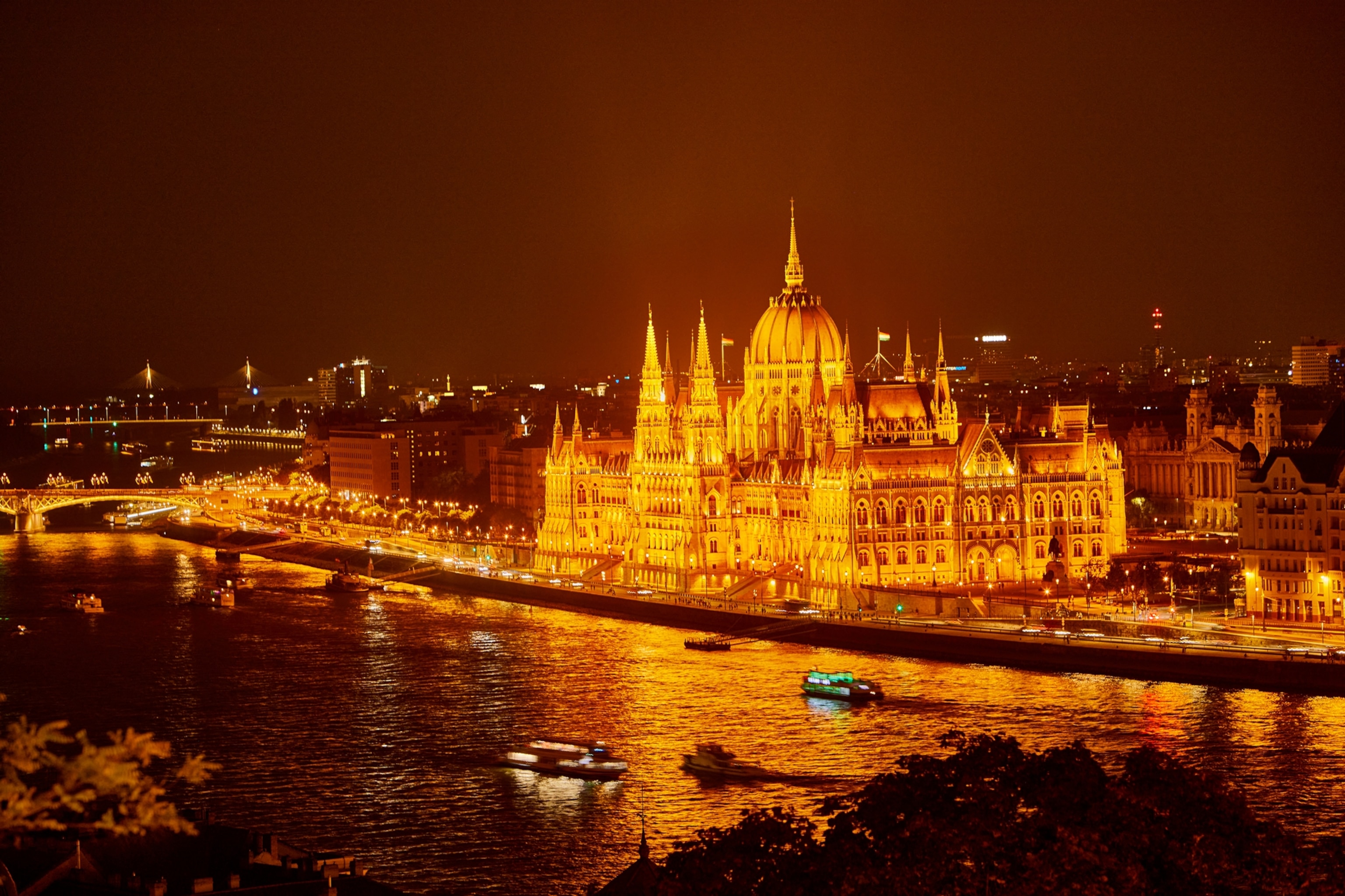 A view of Parliament building in Budapest at nighttime. The glow of the lights is orange and the sky in the background is dark. There are boats in the water.