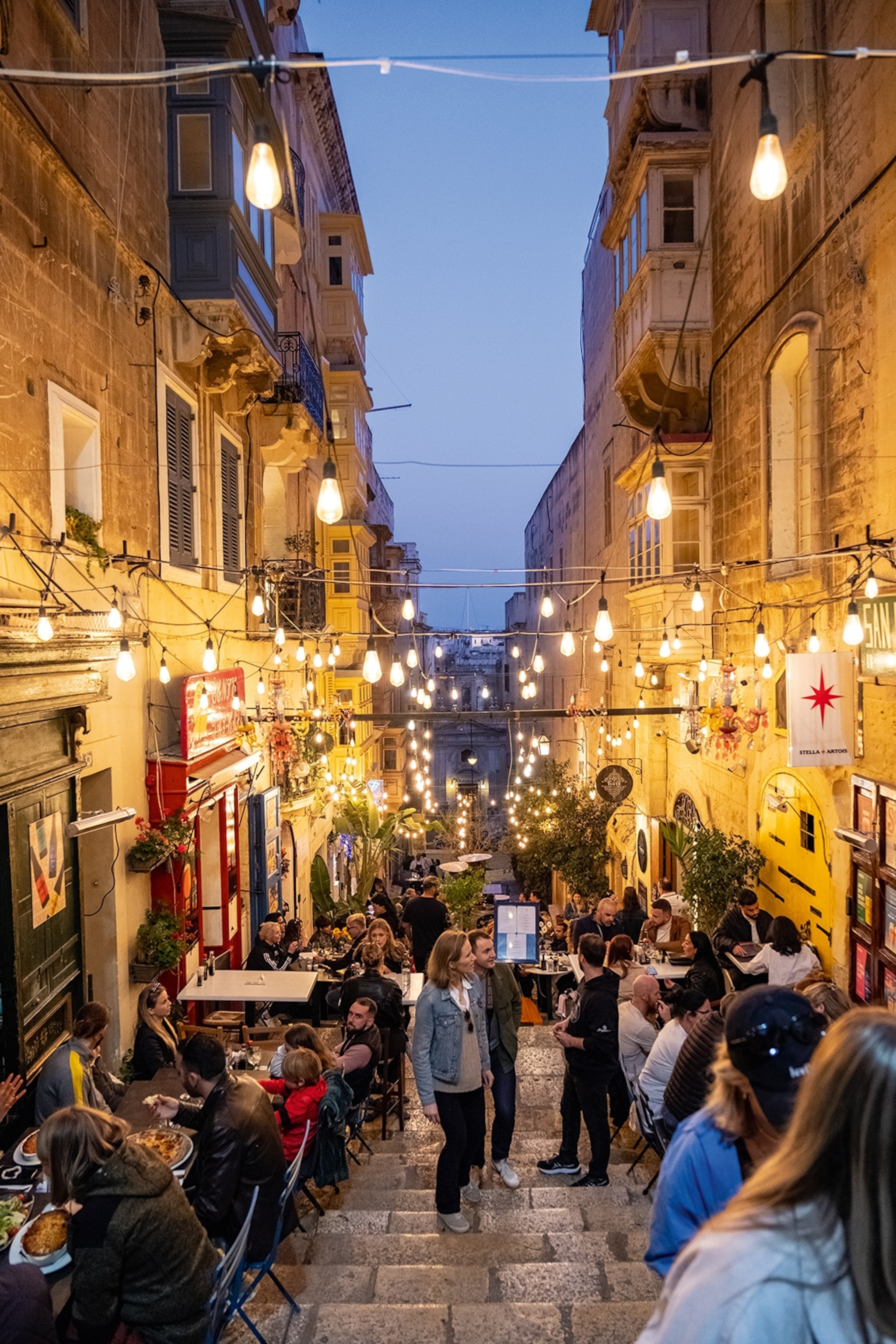 People in an alleyway in Valletta which is populated with bars. Tables line the sides of the steps with people eating and drinking, standing and socialising.
