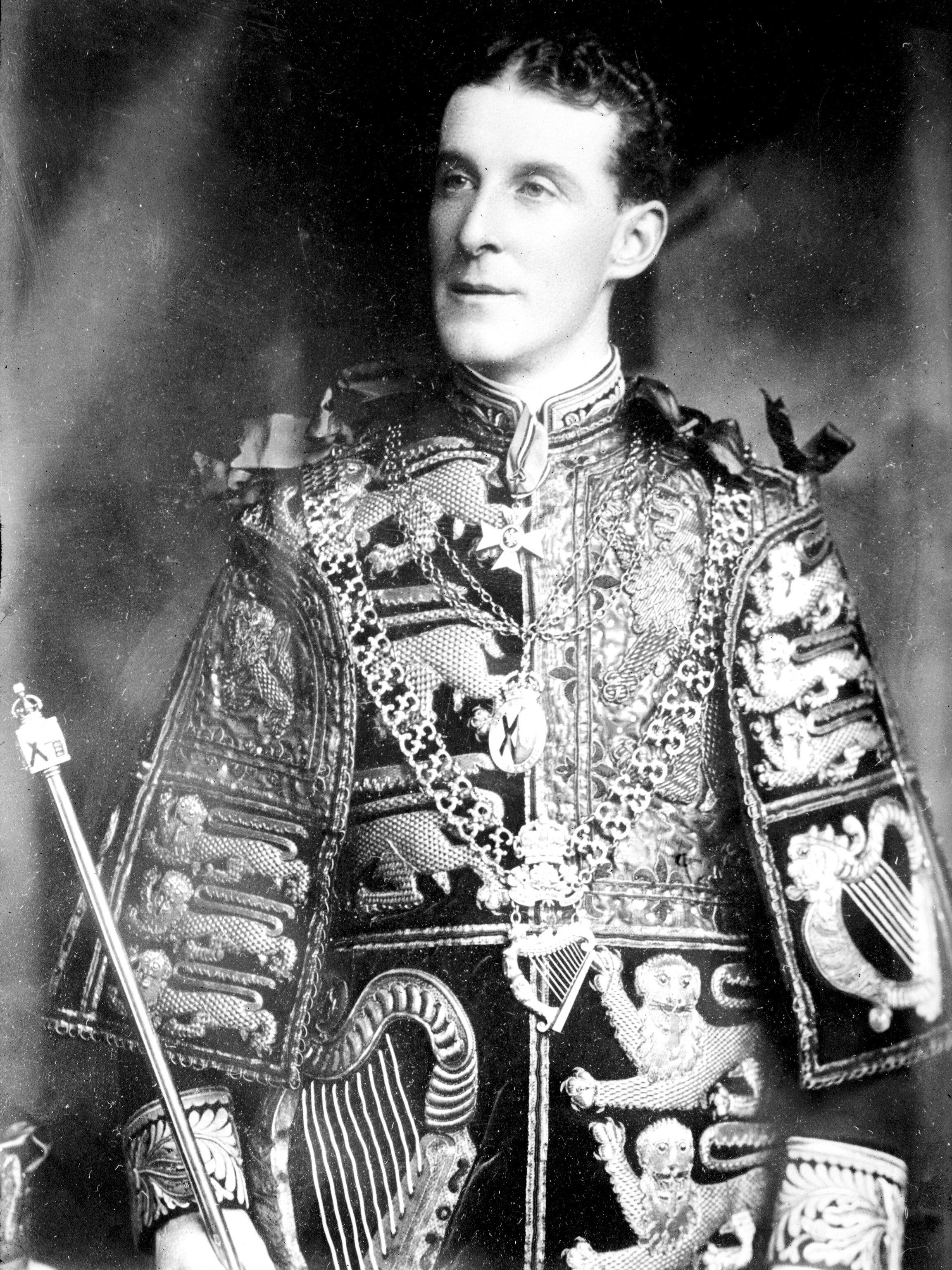 A middle-aged white man holding a scepter poses for a portrait dressed in a ceremonial, embroidered coat and jewelry, in 1902.