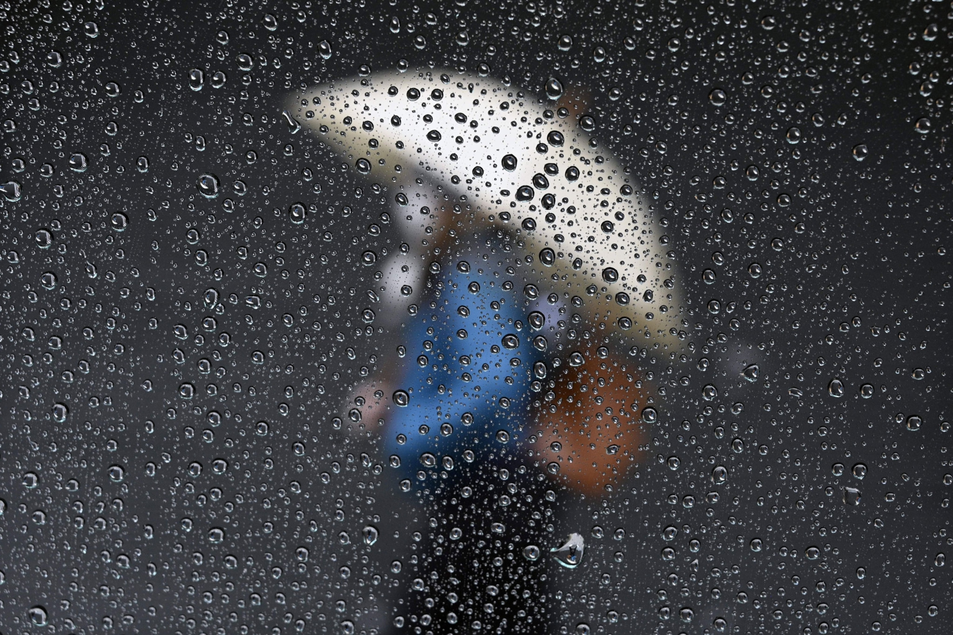 Raindrops on a taxi window as masked people with umbrellas pass by.