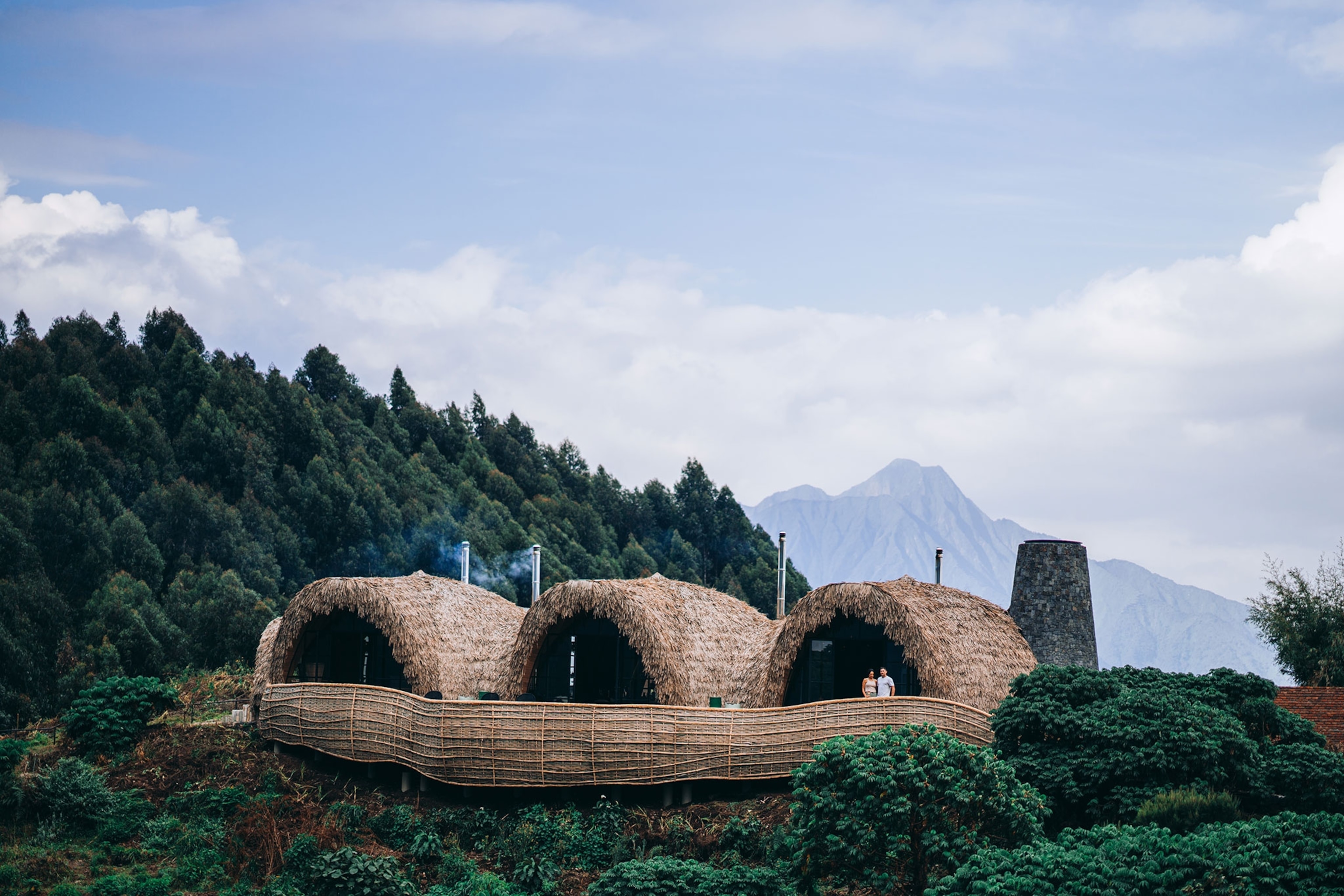 Aerial view of three arch hut-like resort lodges surrounded by jungle mountains and a blue sky above.