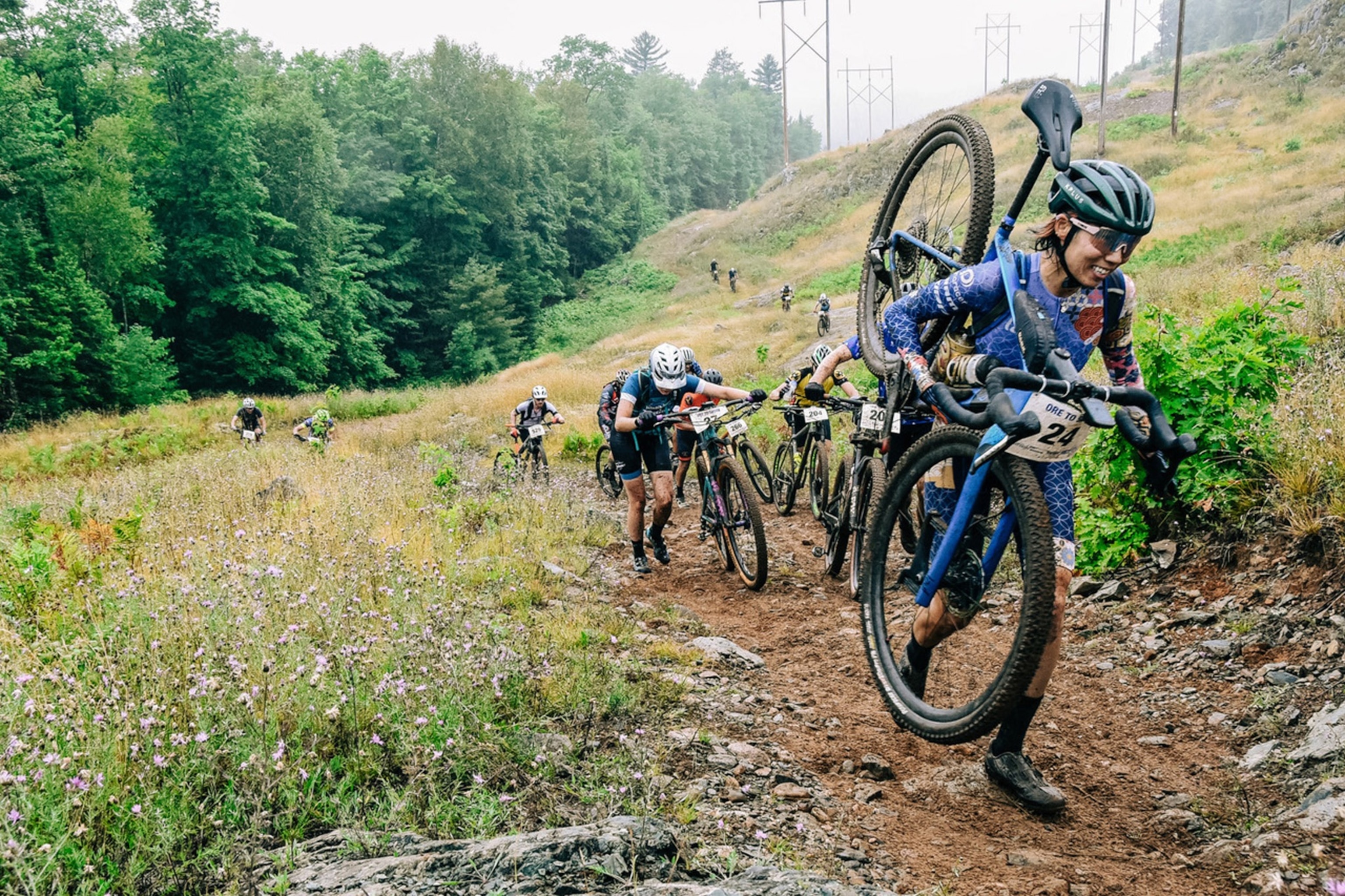A person holds a bike over their shoulder smiling as they tackle a hill in a race.