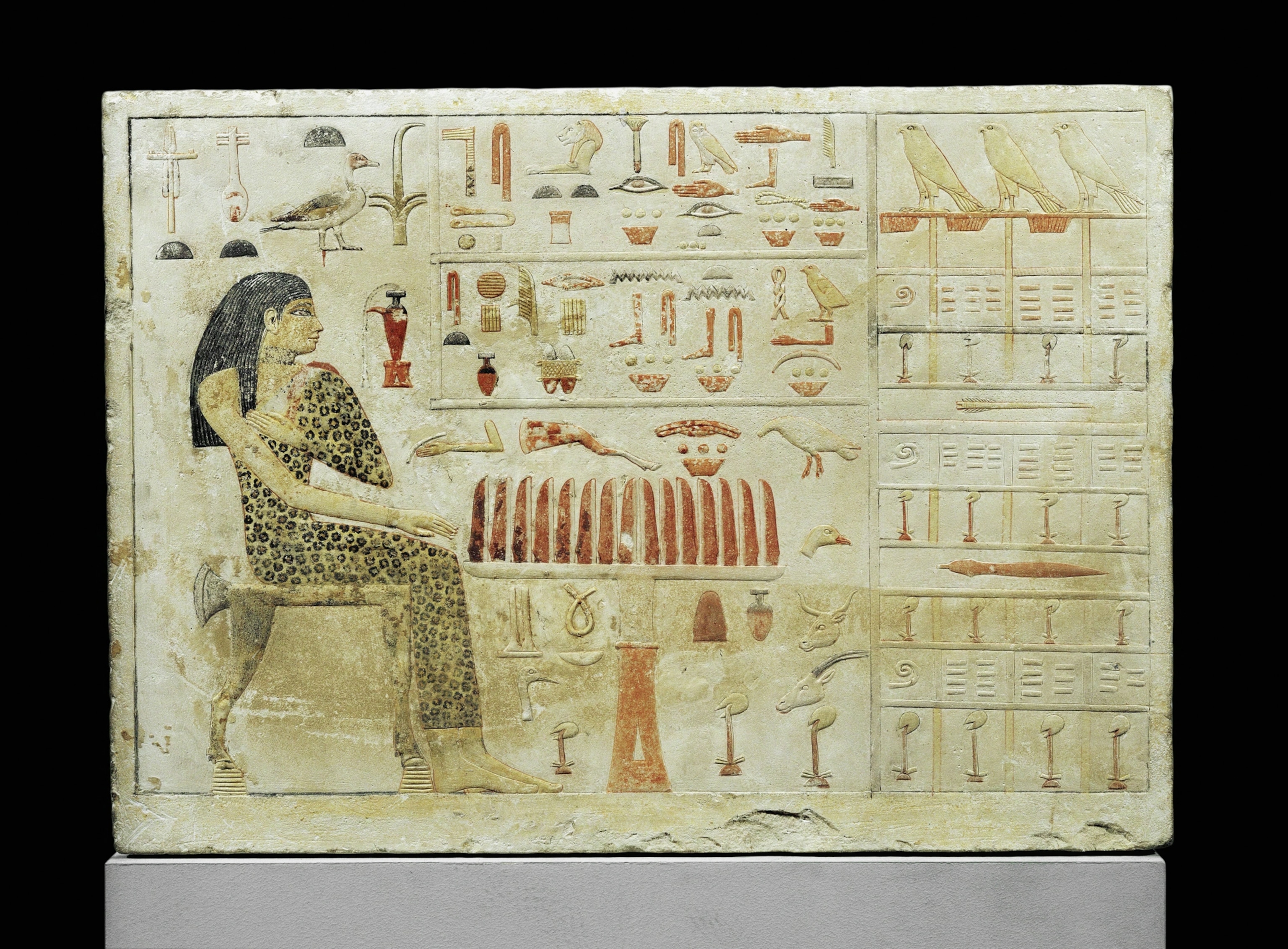 A stone slab shows a woman in leopard skin attire seated at a table. Around her figure are various hieroglyphs.