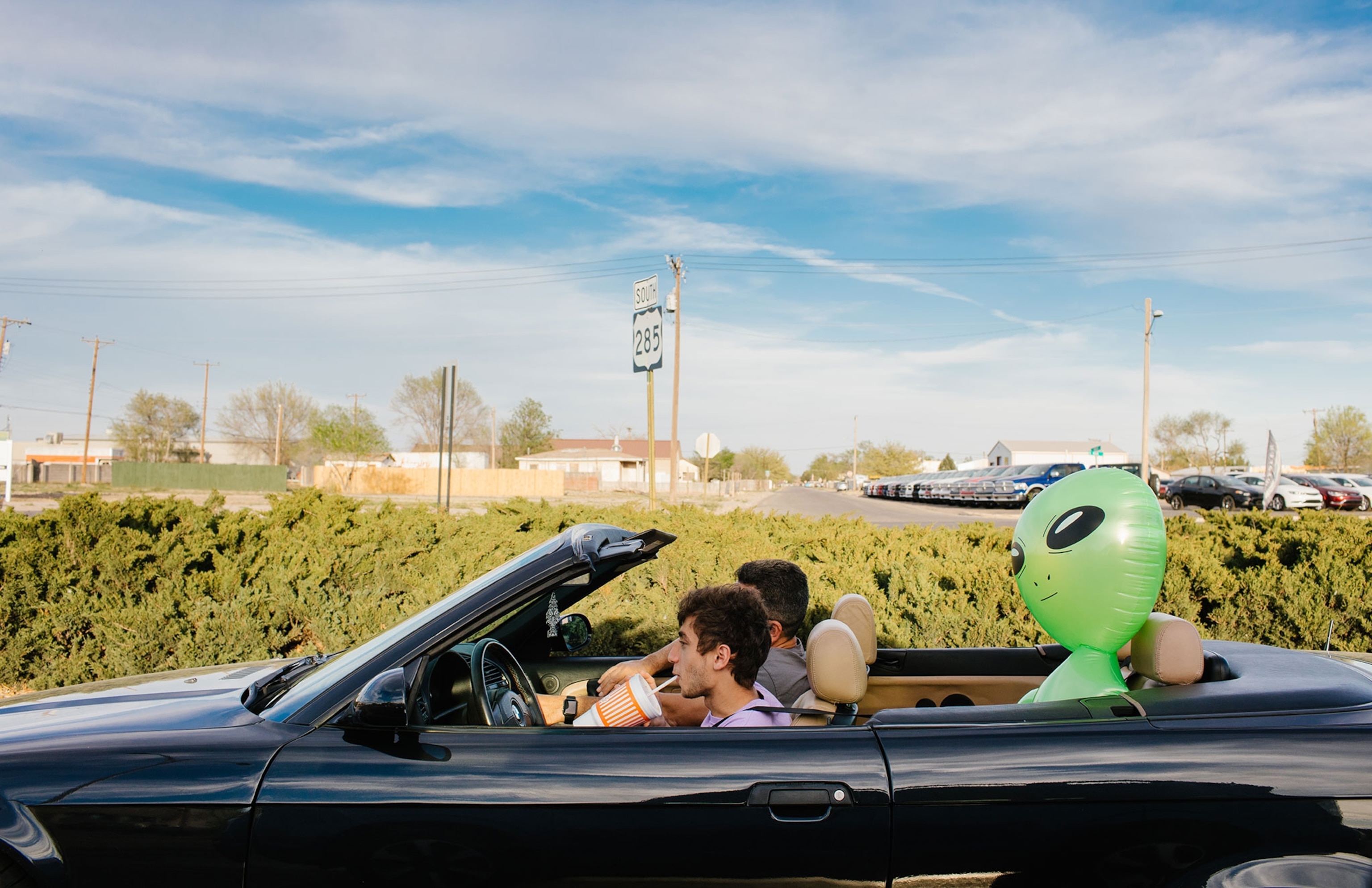 Two people are in a convertible car with a green inflatable alien in the backseat