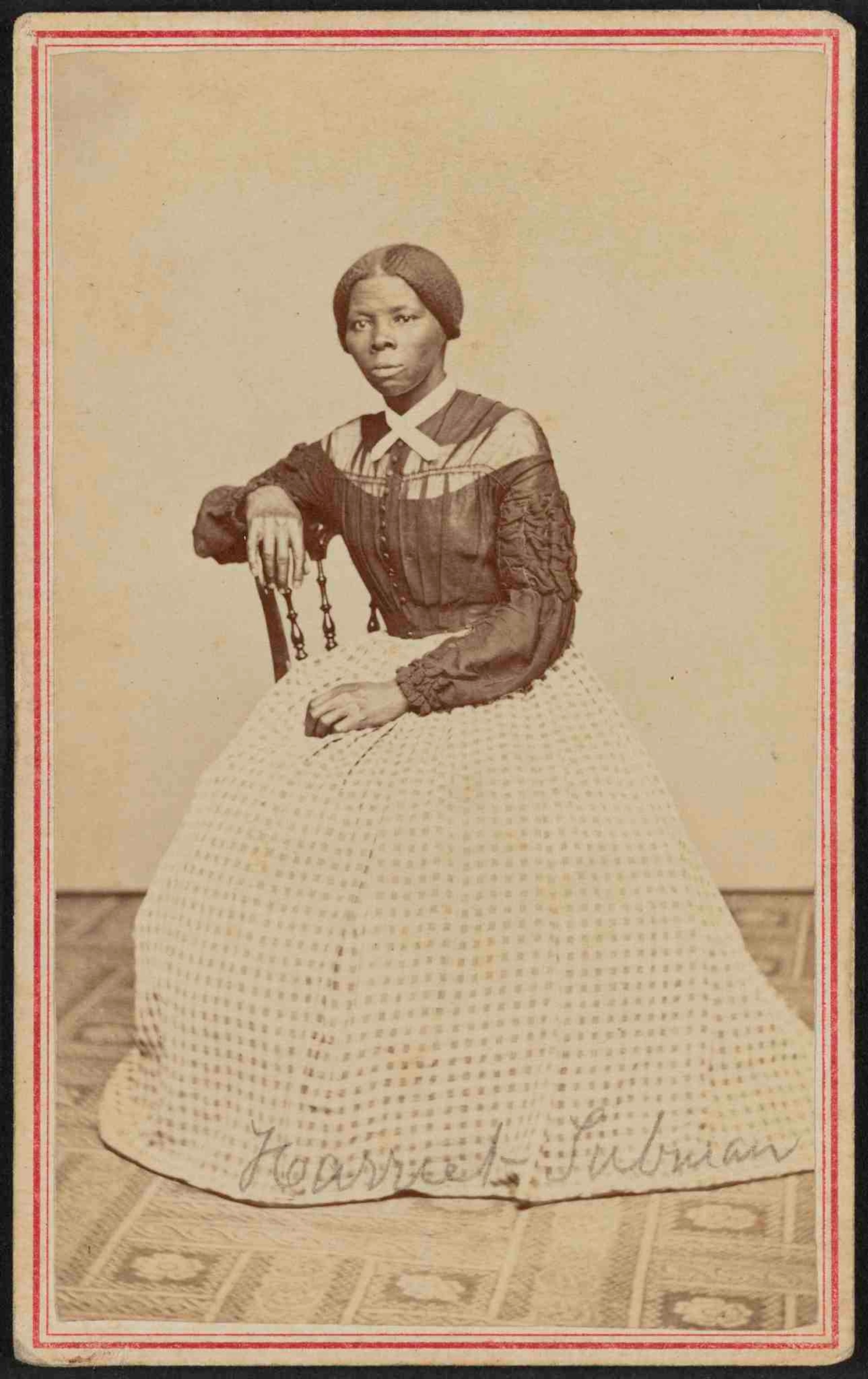 Photograph shows Harriet Tubman at midlife. She is seated, turned toward the left. One hand rests on the back of a wooden chair, another rests in her lap.