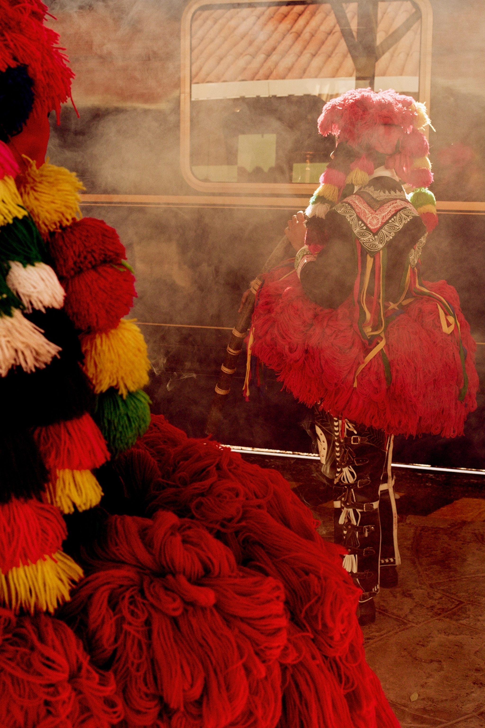 Two dancers on a cloudy, hot train platform, dressed in celebratory costumes of multi-coloured pom-poms.