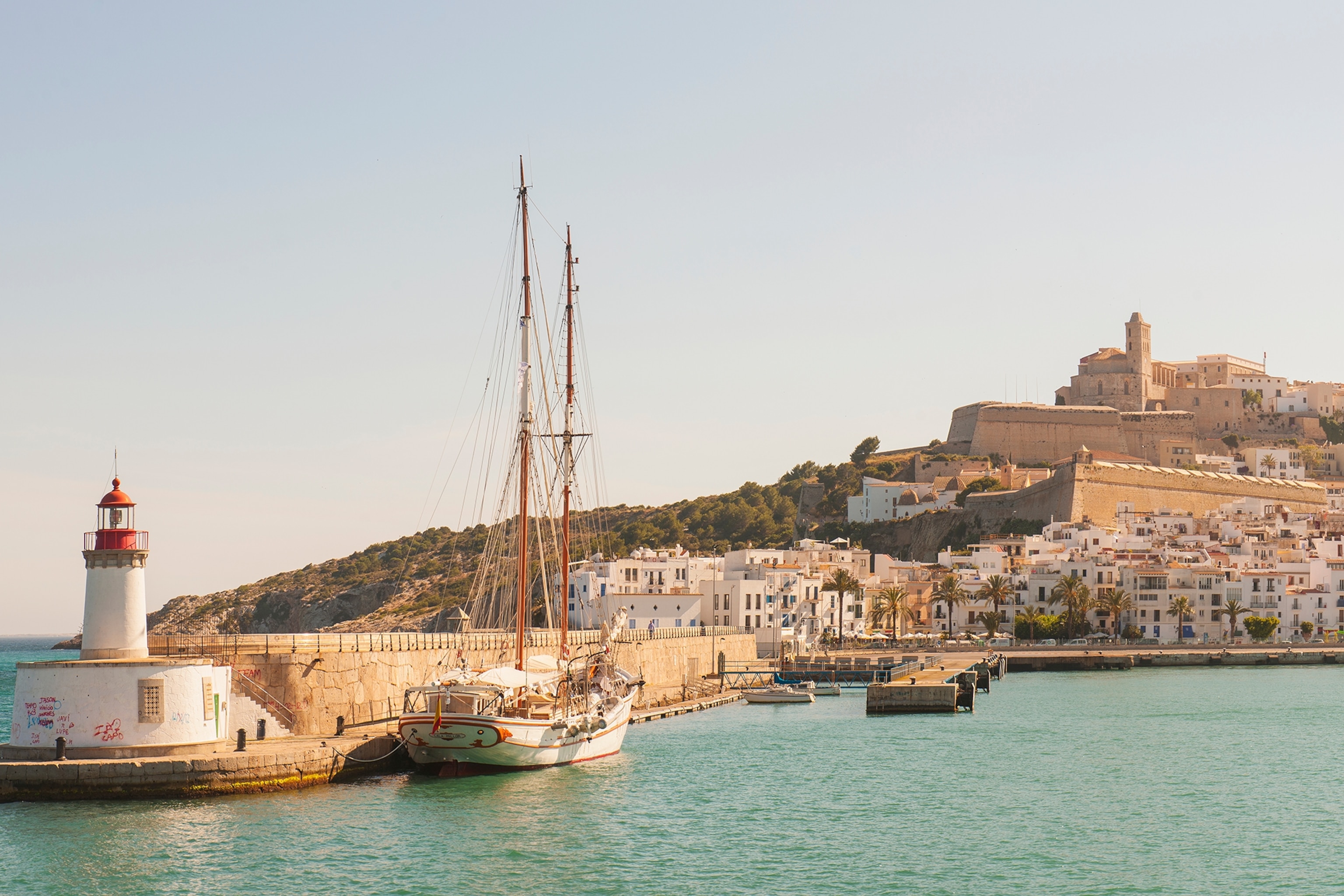 A seaside view on the hill-positioned Ibiza Town with a sailing boat and small lighthouse in the left hand foreground.