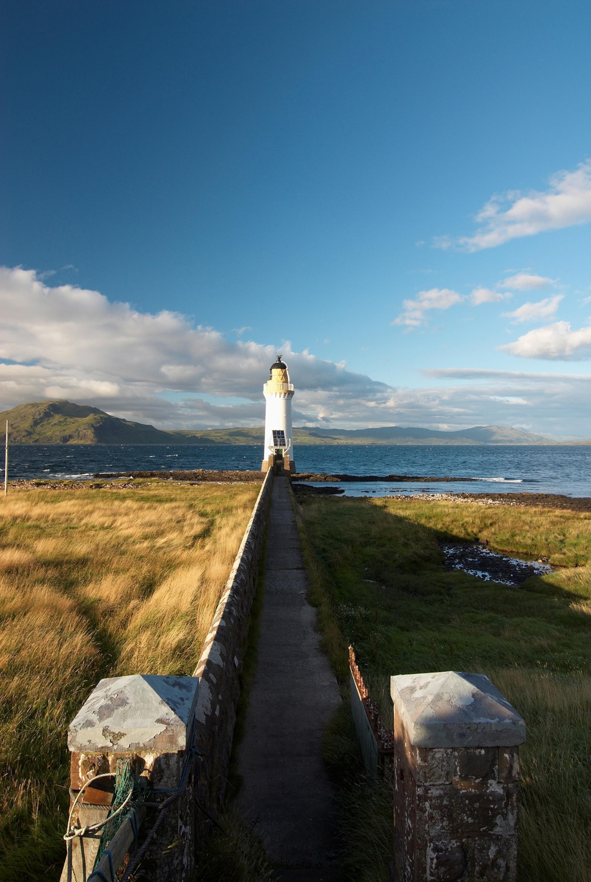 A light house sits at the end of a long walkway with brush around it under a deep blue sky.