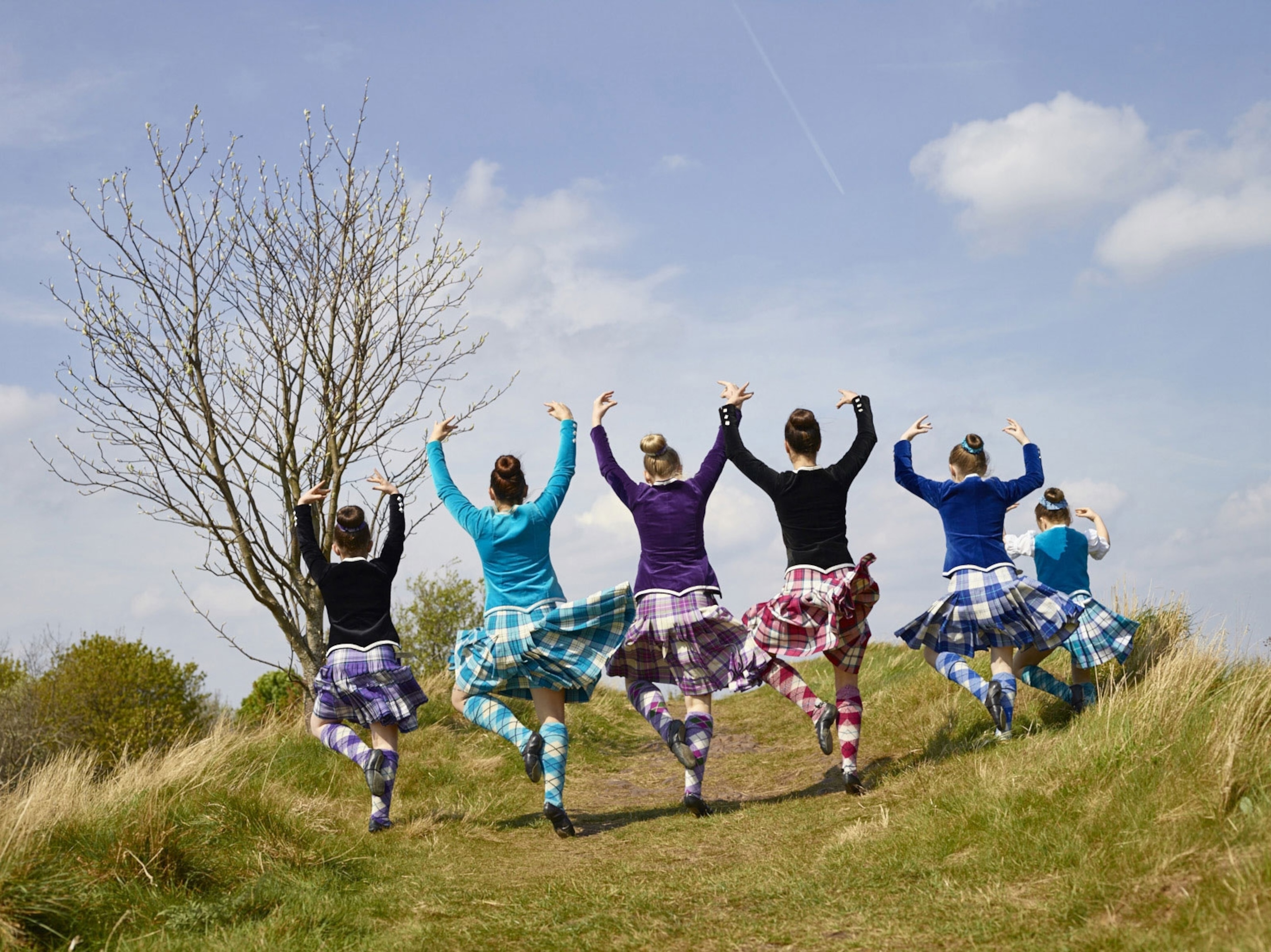 A group of girls jump in unison with arms raised as part of a Highland dance on a grassy hill.