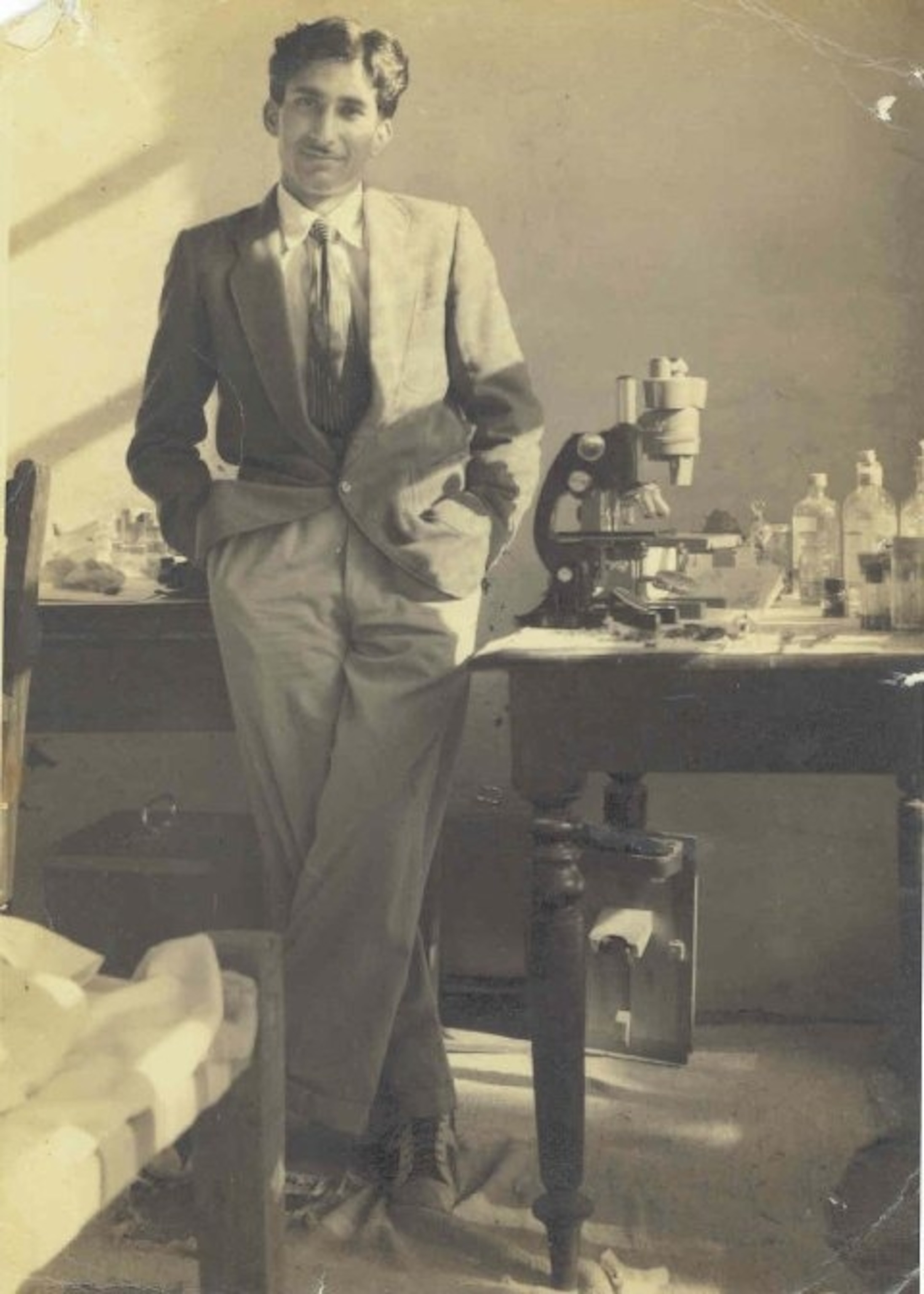 black and white photo of man in suit standing against desk with microscope
