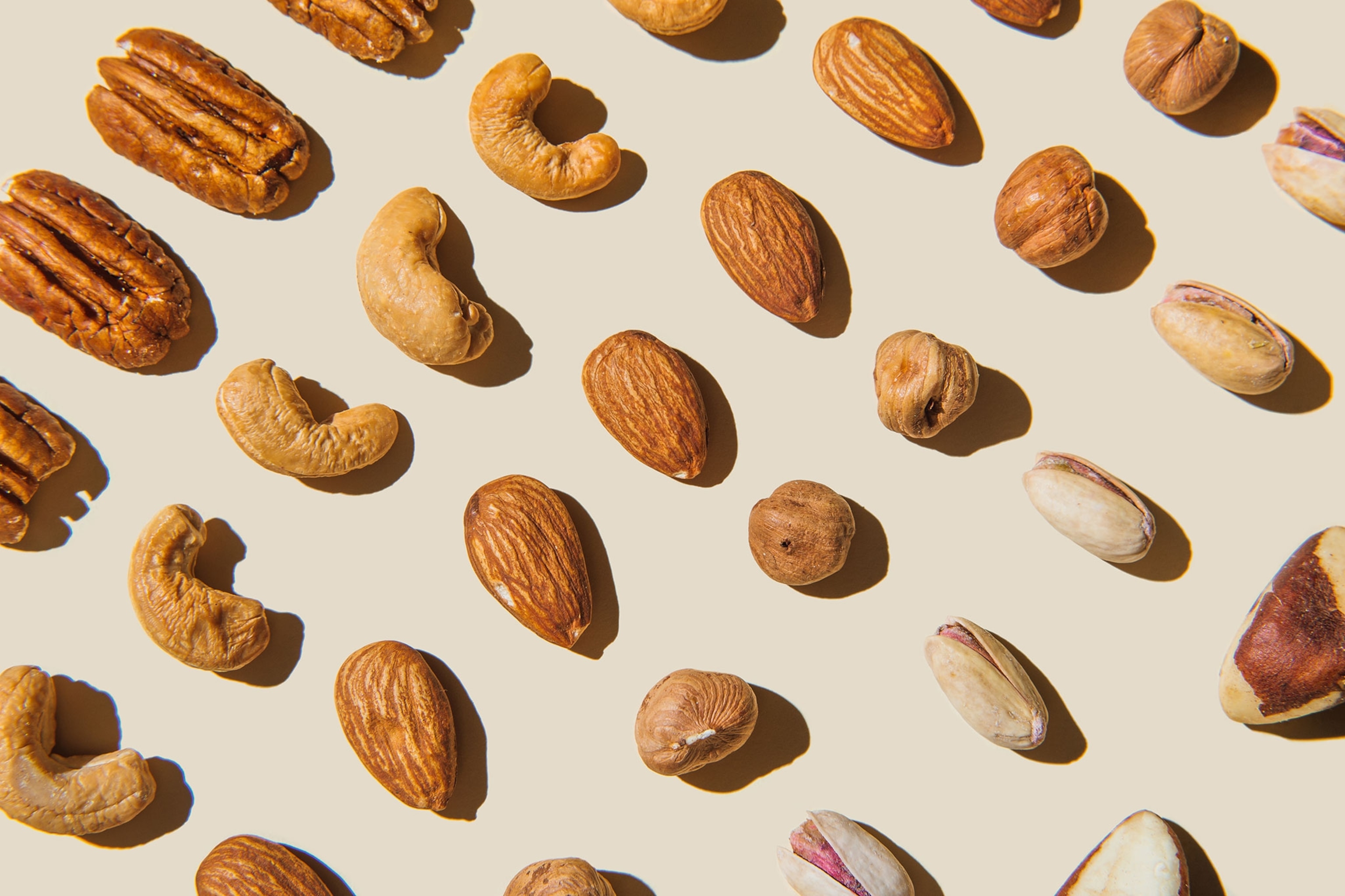 Pecans, brazil nuts, almonds, hazelnuts, pistachios, and cashews lined in rows on a tan colored background.