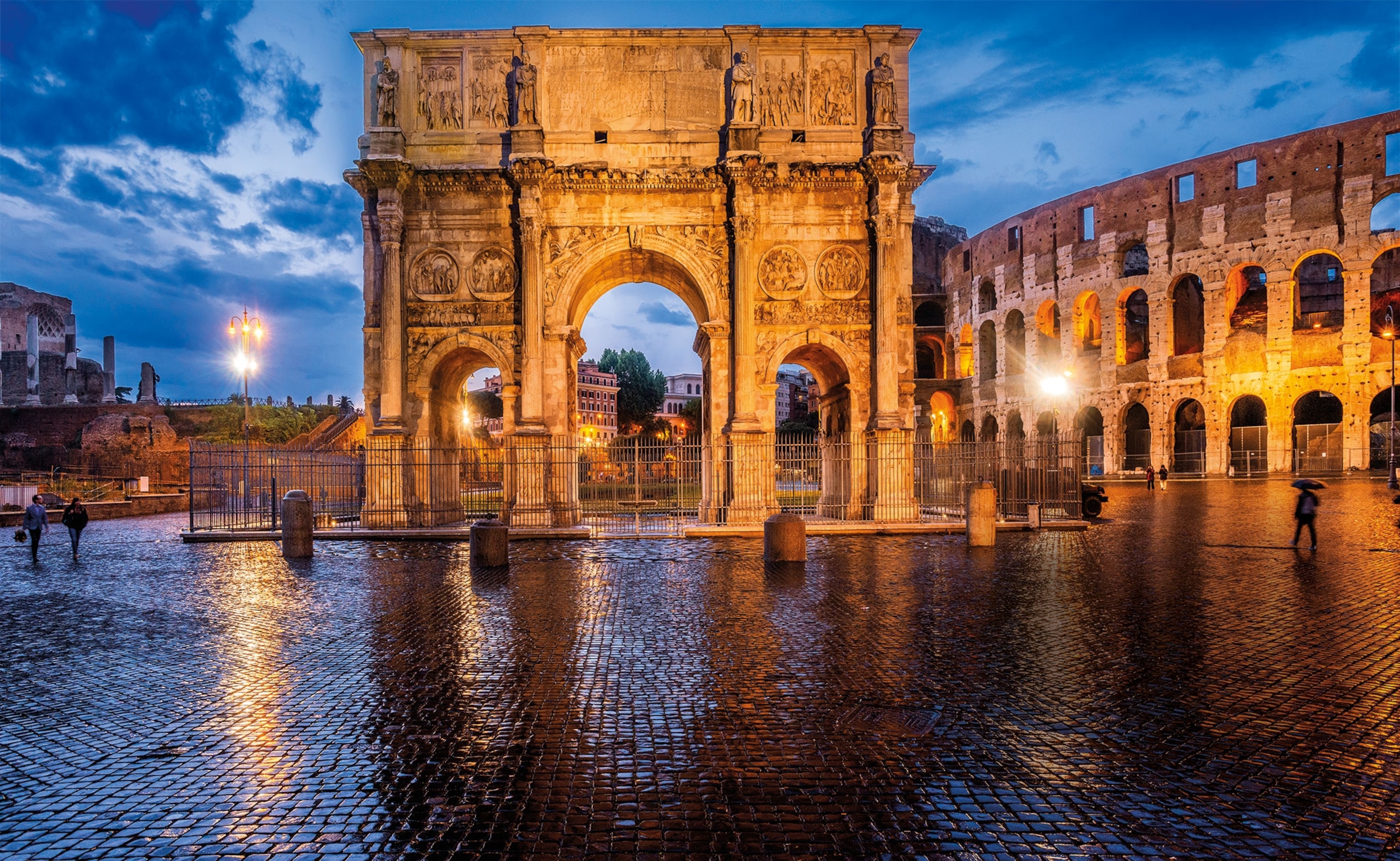 A marble arch with carvings of various scenes is shown lit up at night next to the Colosseum