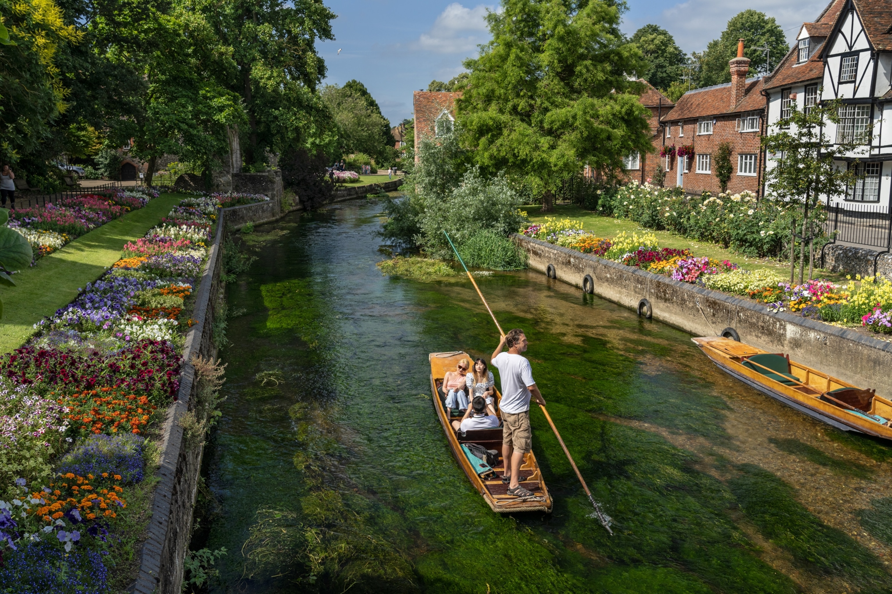 a person is standing on a little wooden boat with a large wooden pole while three people are seated enjoying the scenic view of flowers and architecture.