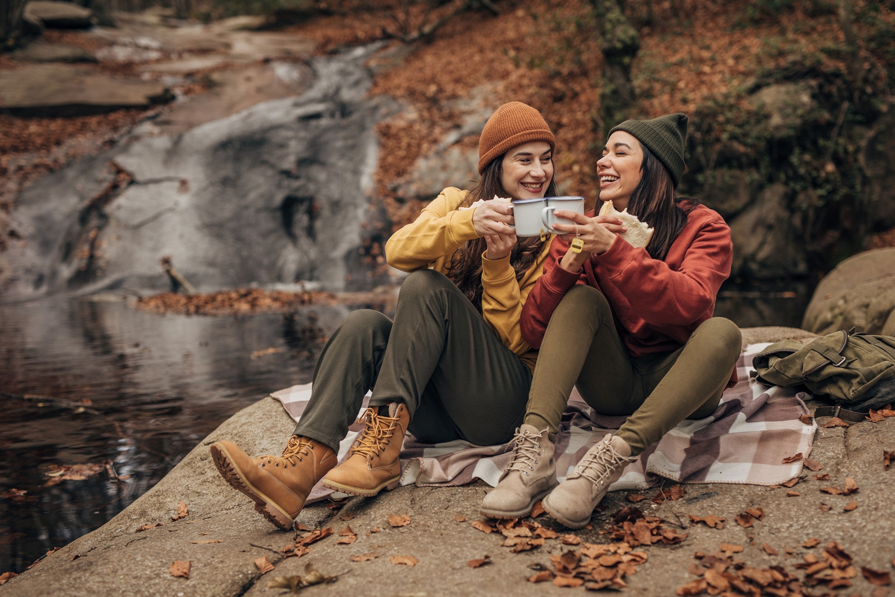 Two women in fall attire sitting in the woods nears water.