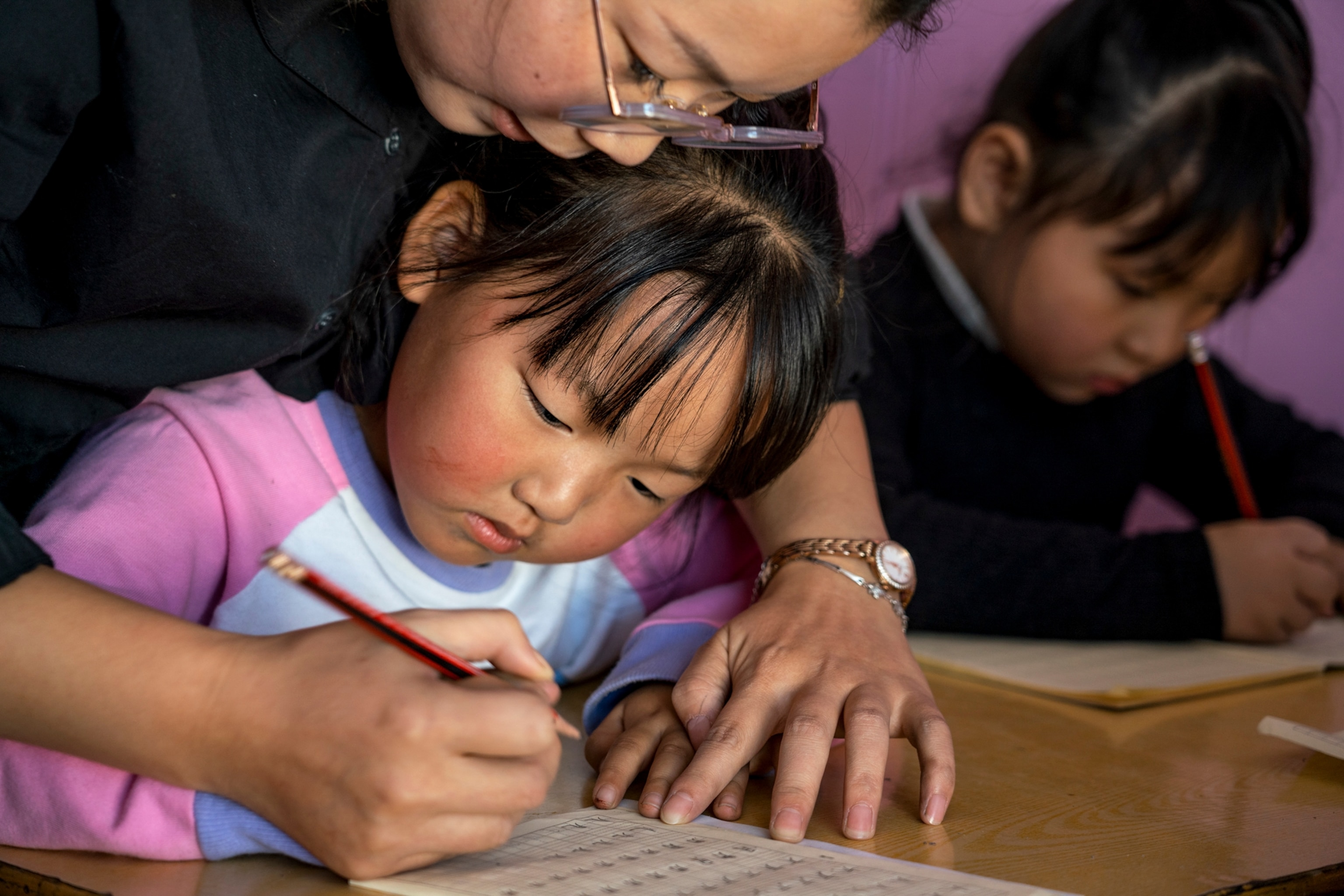 Picture of a little girl at a desk with an adult helping her learn to write on paper with a pencil.