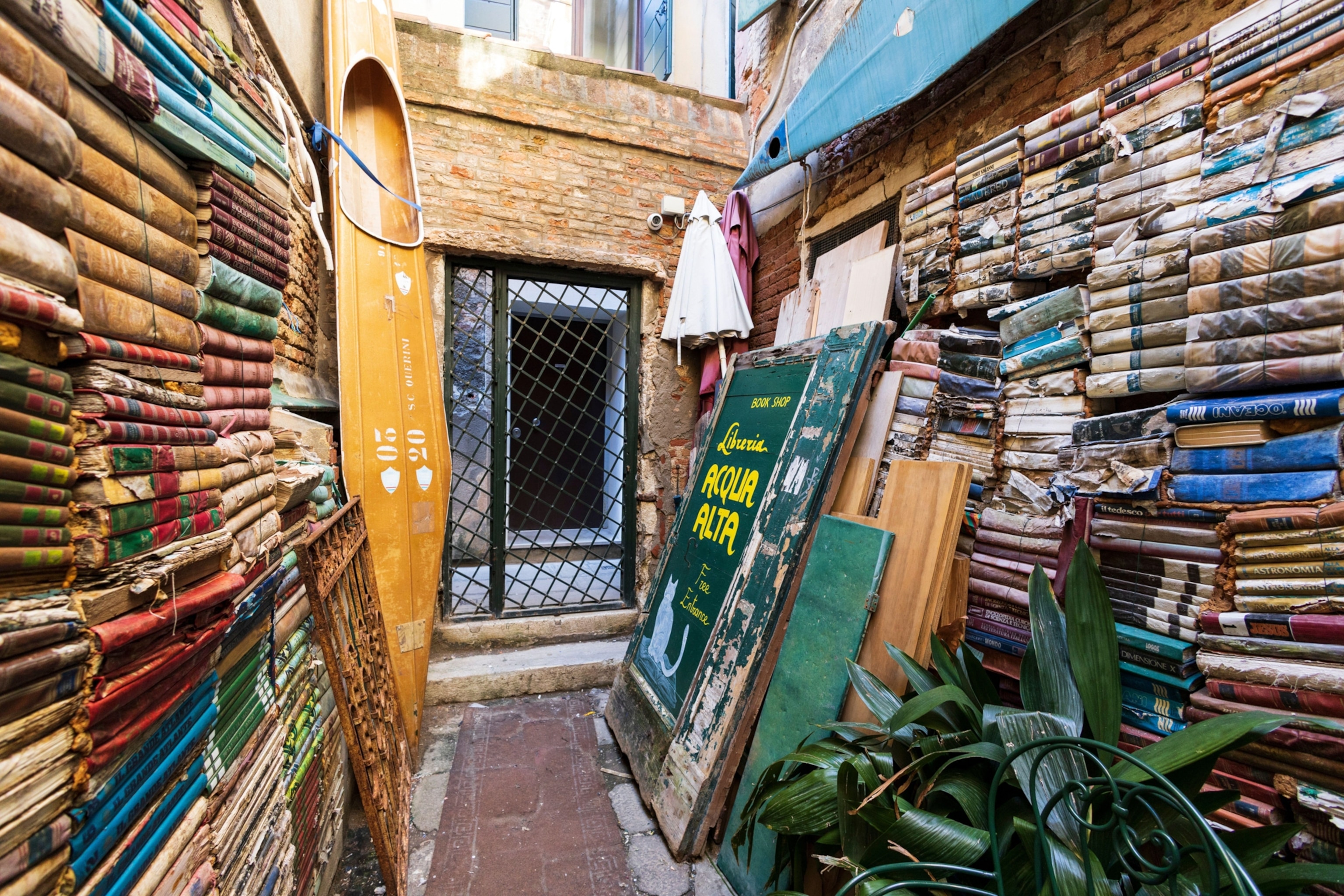 Stacks of old books lining a narrow stone alley. At the end of the short alley, an old gondola stands beside a closed metal gate.