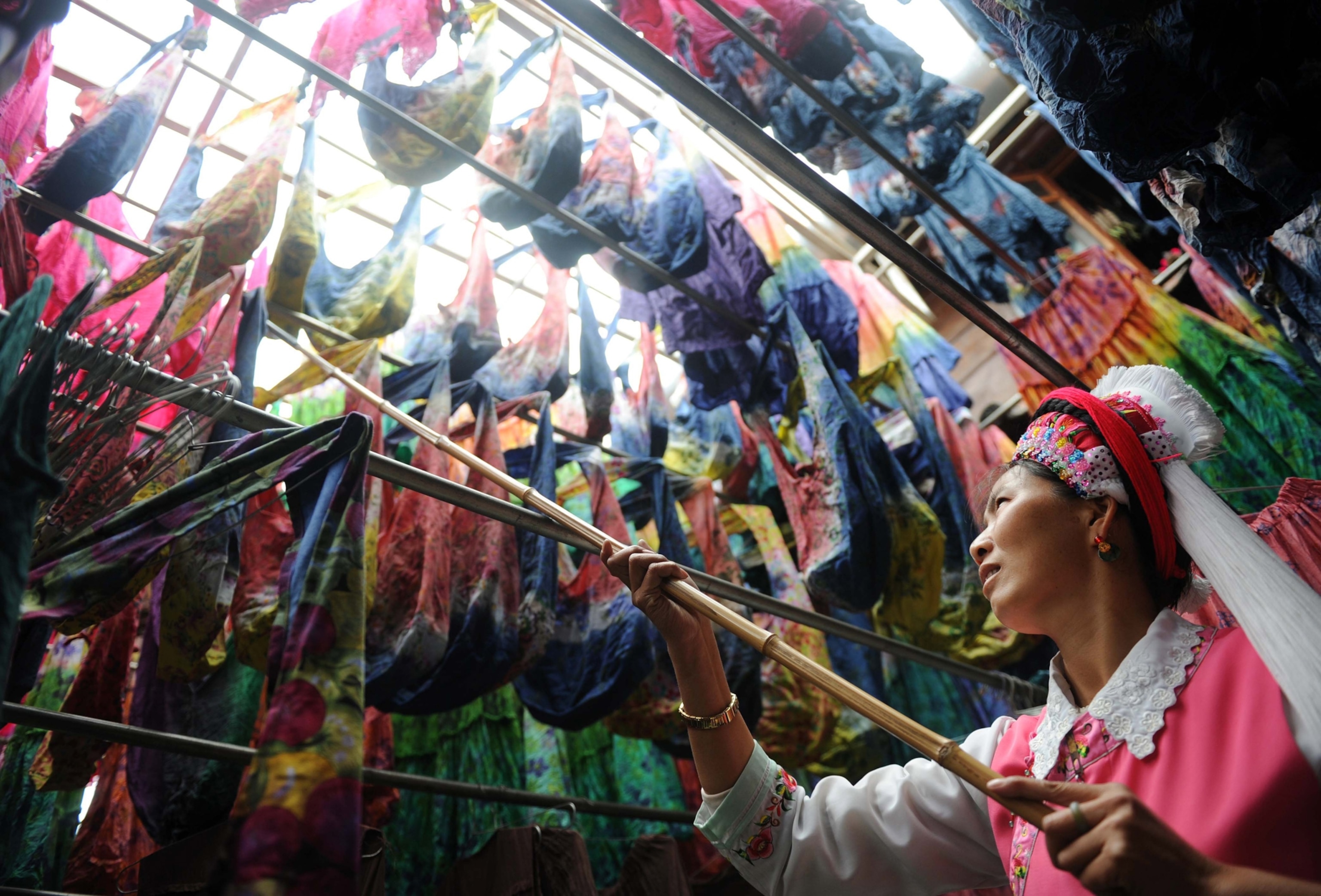 A woman of the Bai ethnic group uses a bamboo pole to adjust colorfully dyed clothes drying along a series of bamboo poles above.