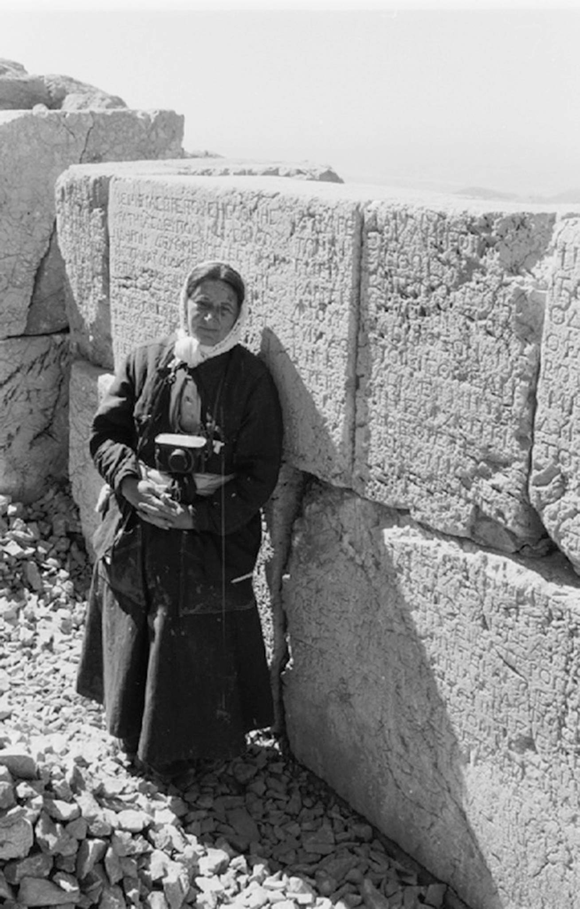 A woman in dark clothes and a scarf around her head stands with a camera in front of stone blocks with engraved words