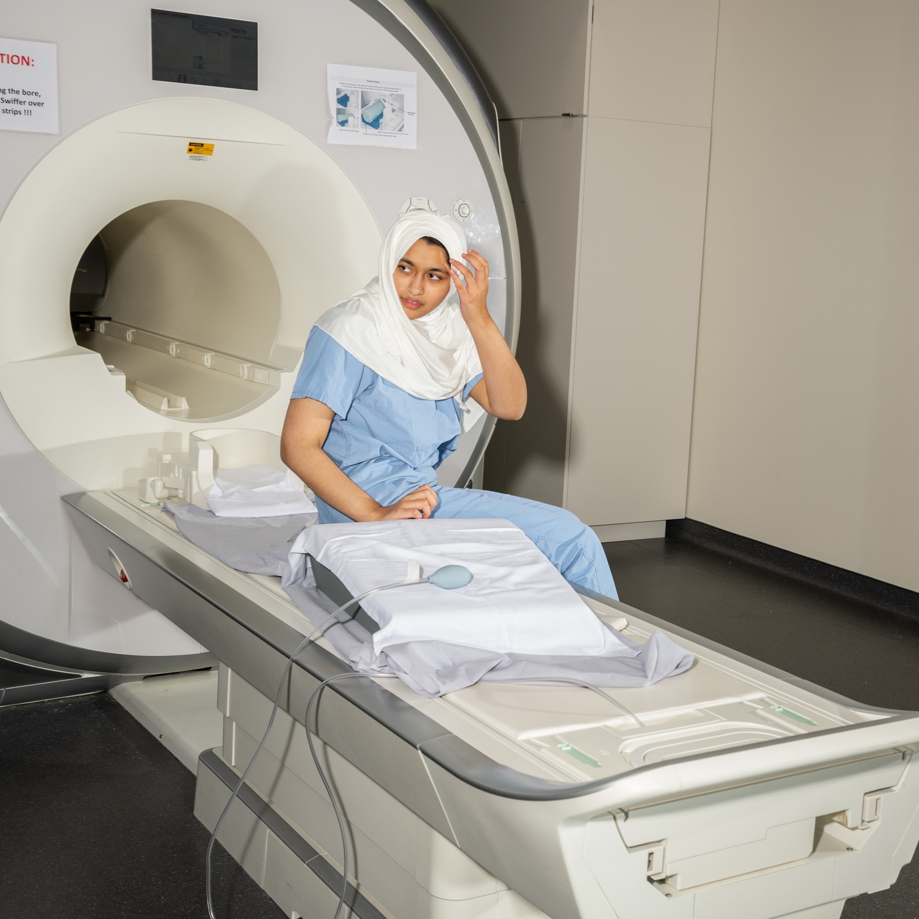 A teen girl wearing a white hijab and blue scrubs sits on an MRI table.